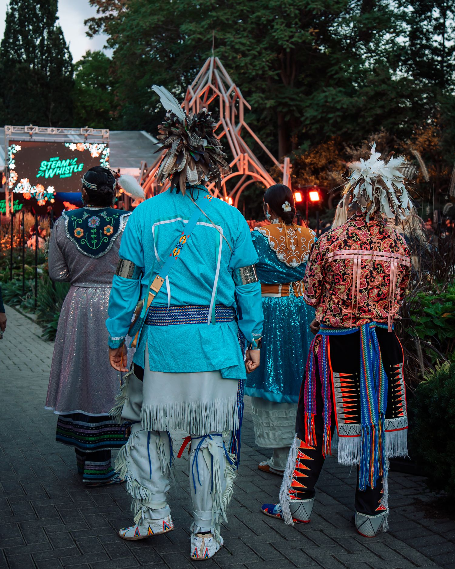Three Indigenous people in traditional regalia with feathered headdresses walk together at an outdoor festival at dusk.
