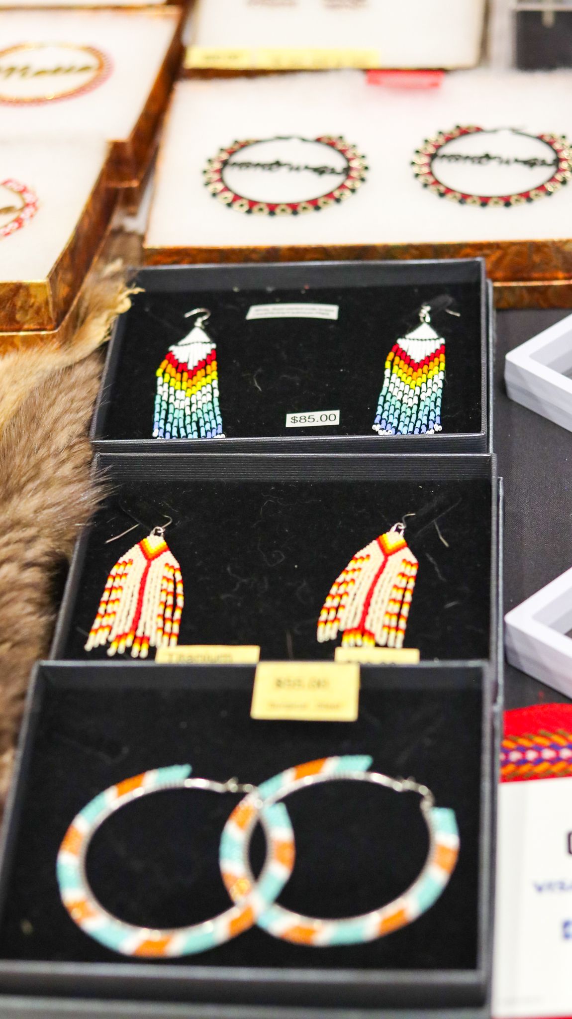 Colorful Native American beaded earrings displayed in black jewelry boxes, featuring fringe and hoop styles with vibrant patterns.