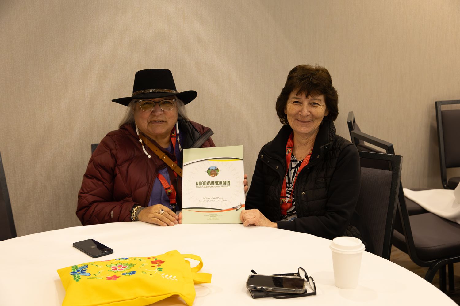 Two people sit at round table holding certificate at indoor Indigenous community gathering.