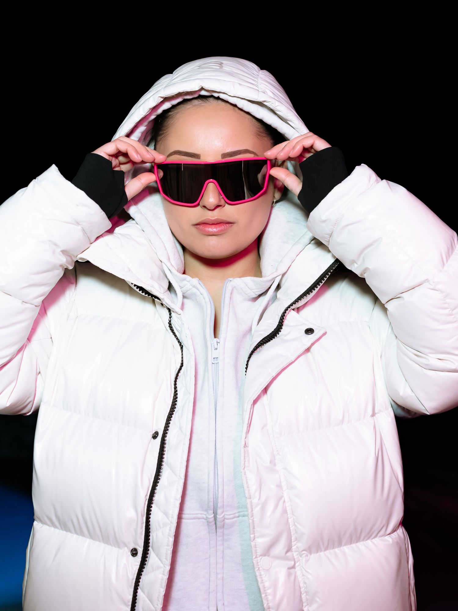 Woman in white hooded puffer jacket adjusting pink shield sunglasses against a dark background.