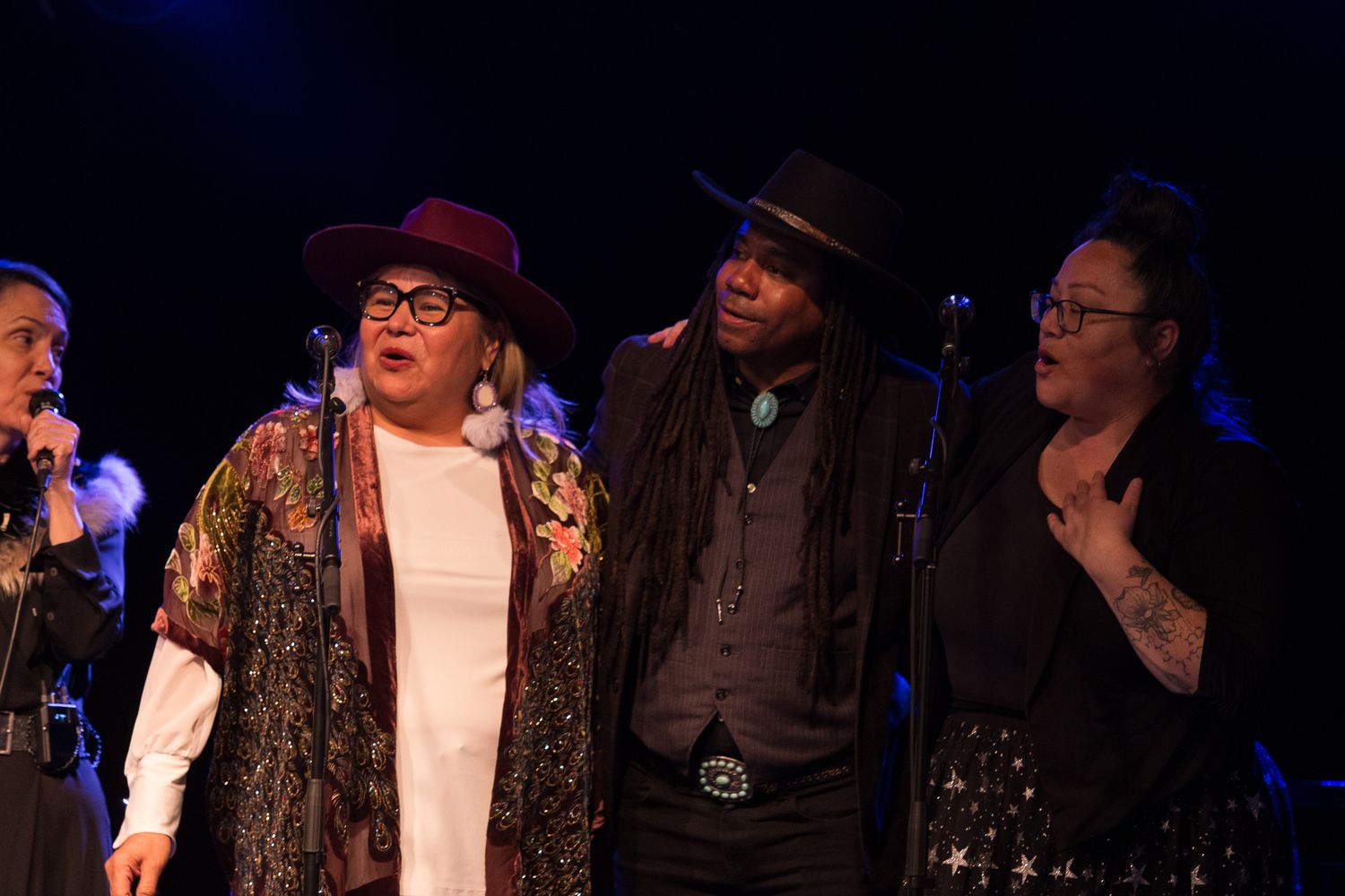 Three performers sing together on stage, wearing stylish hats and colorful outfits under blue stage lighting.