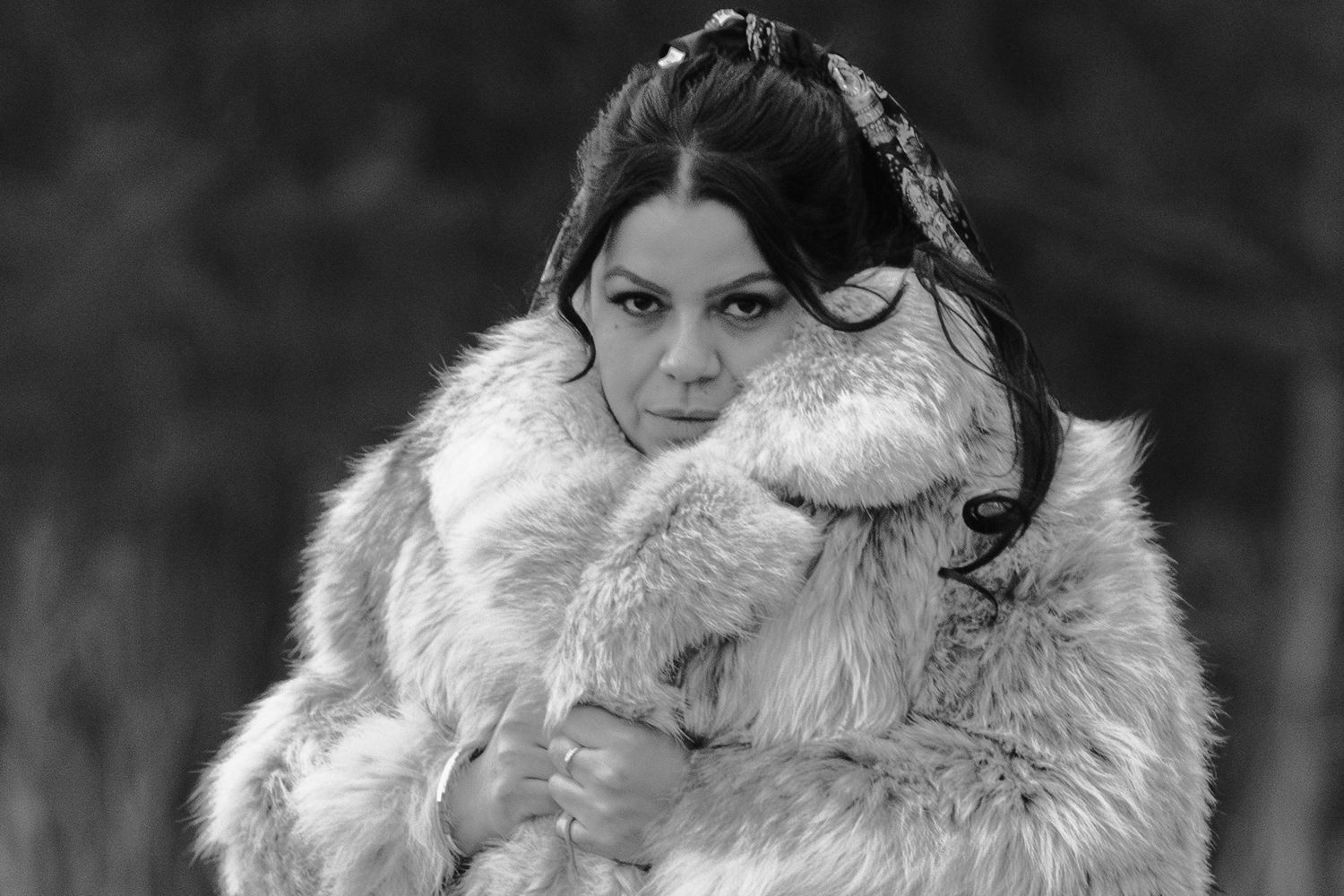 Black and white portrait of a young woman wrapped in a large fur coat, gazing at the camera outdoors.