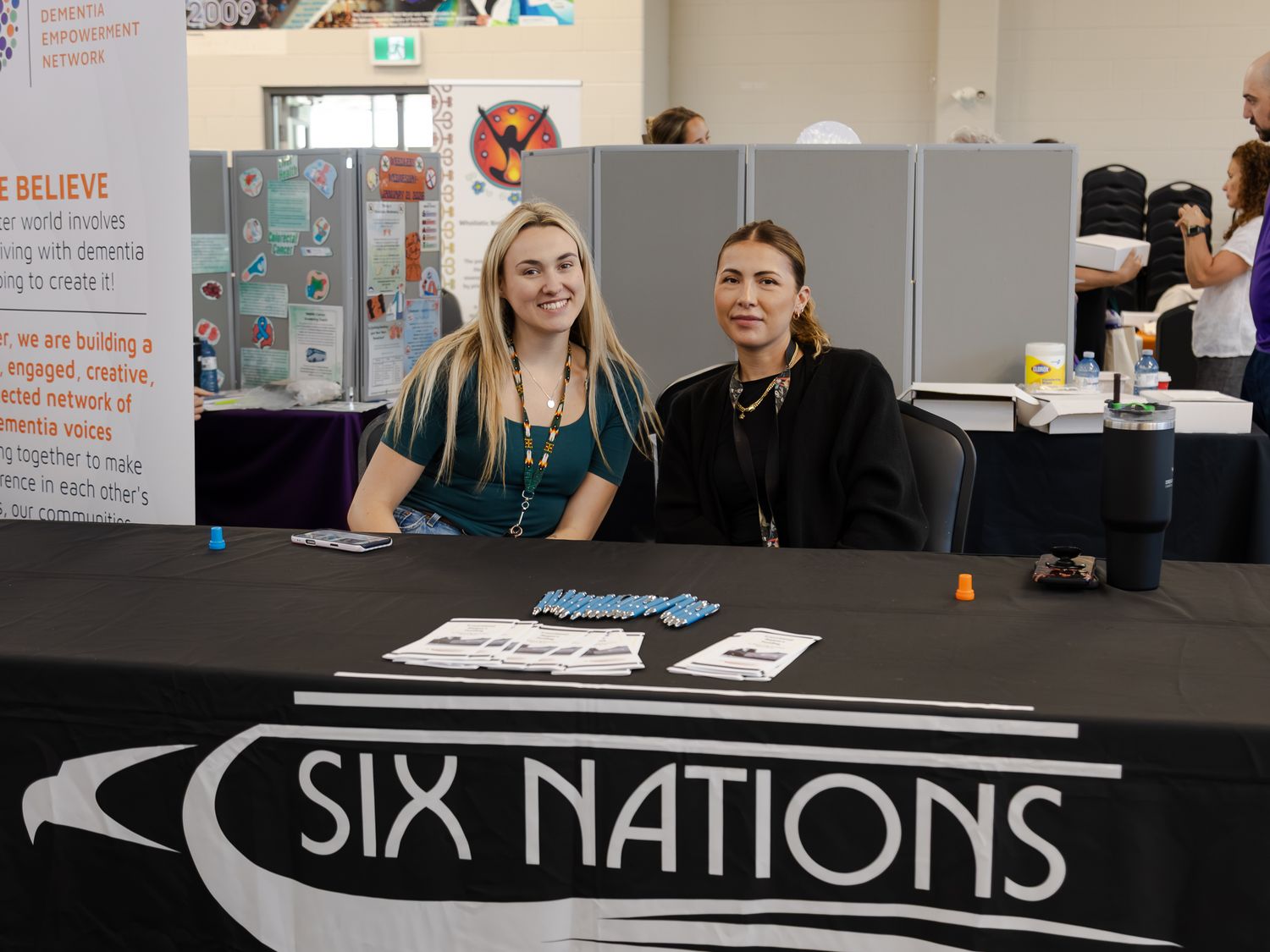 Two women smile at Six Nations information booth table at a dementia awareness conference exhibition hall.