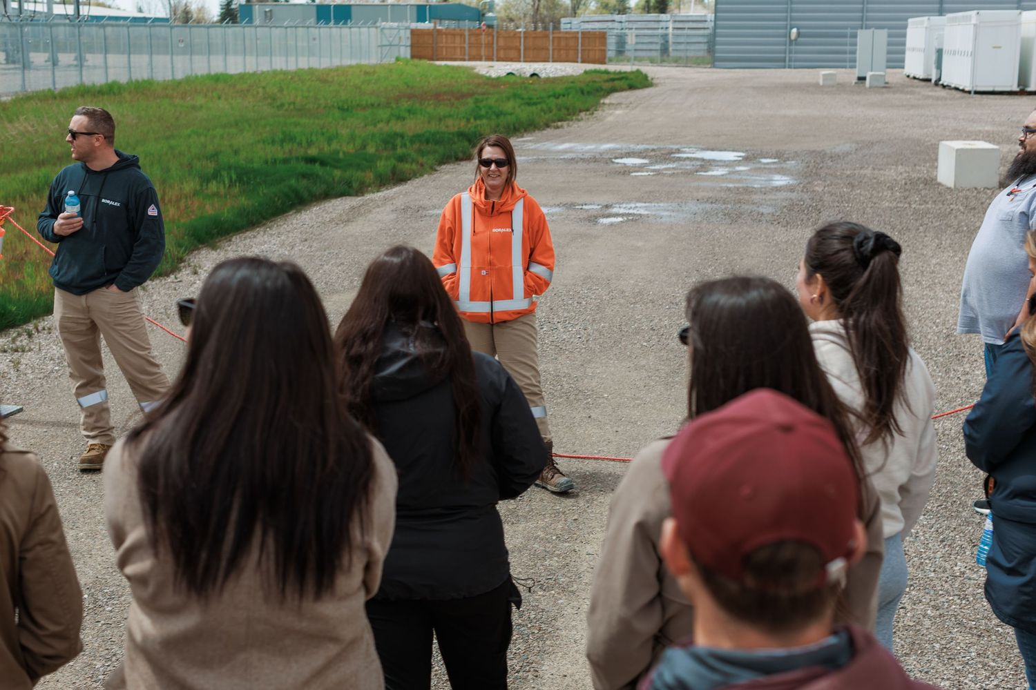 Worker in orange safety vest addresses group of students outdoors at industrial facility.