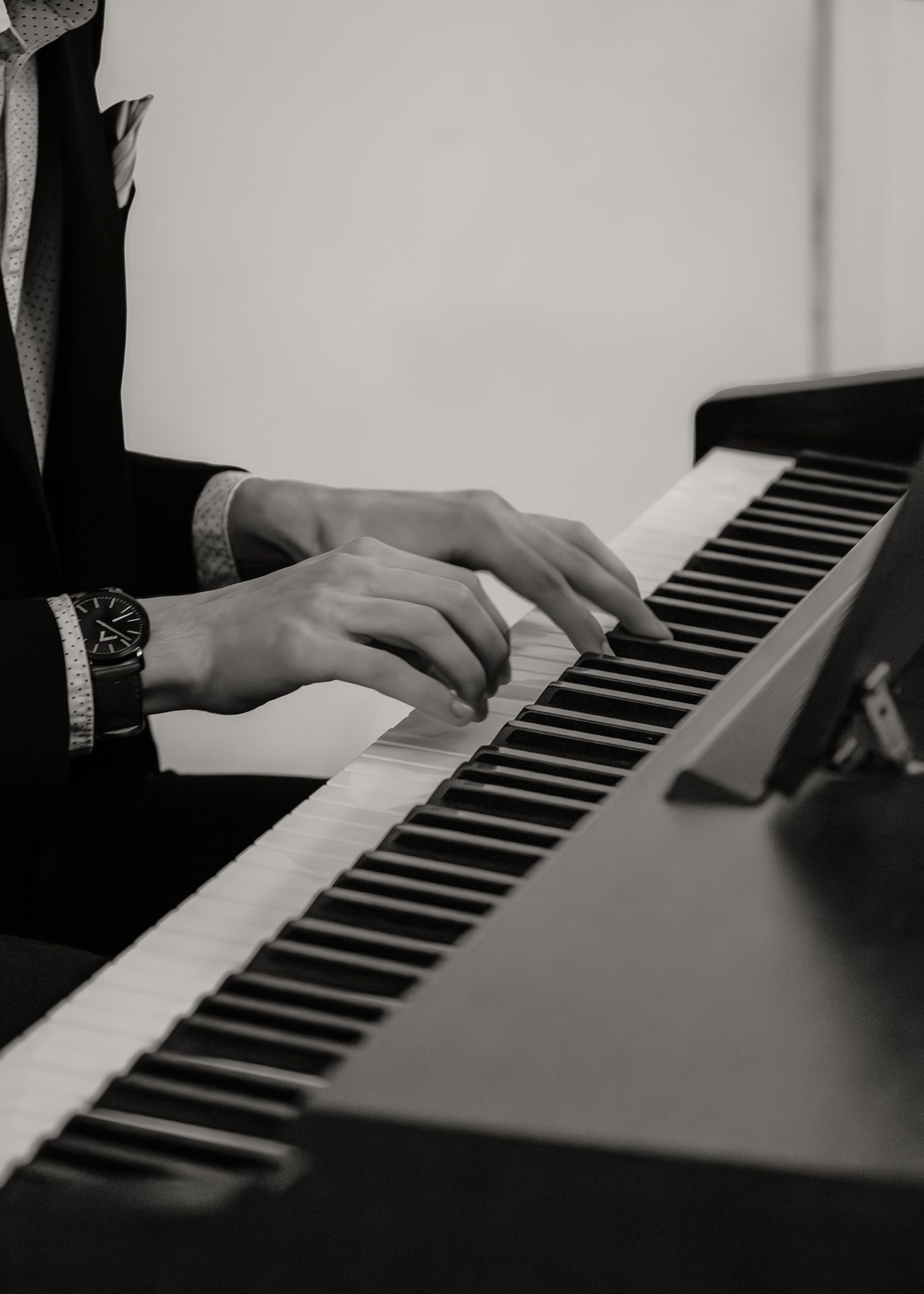 Close-up of hands playing piano keys in black and white, wearing a suit and watch.