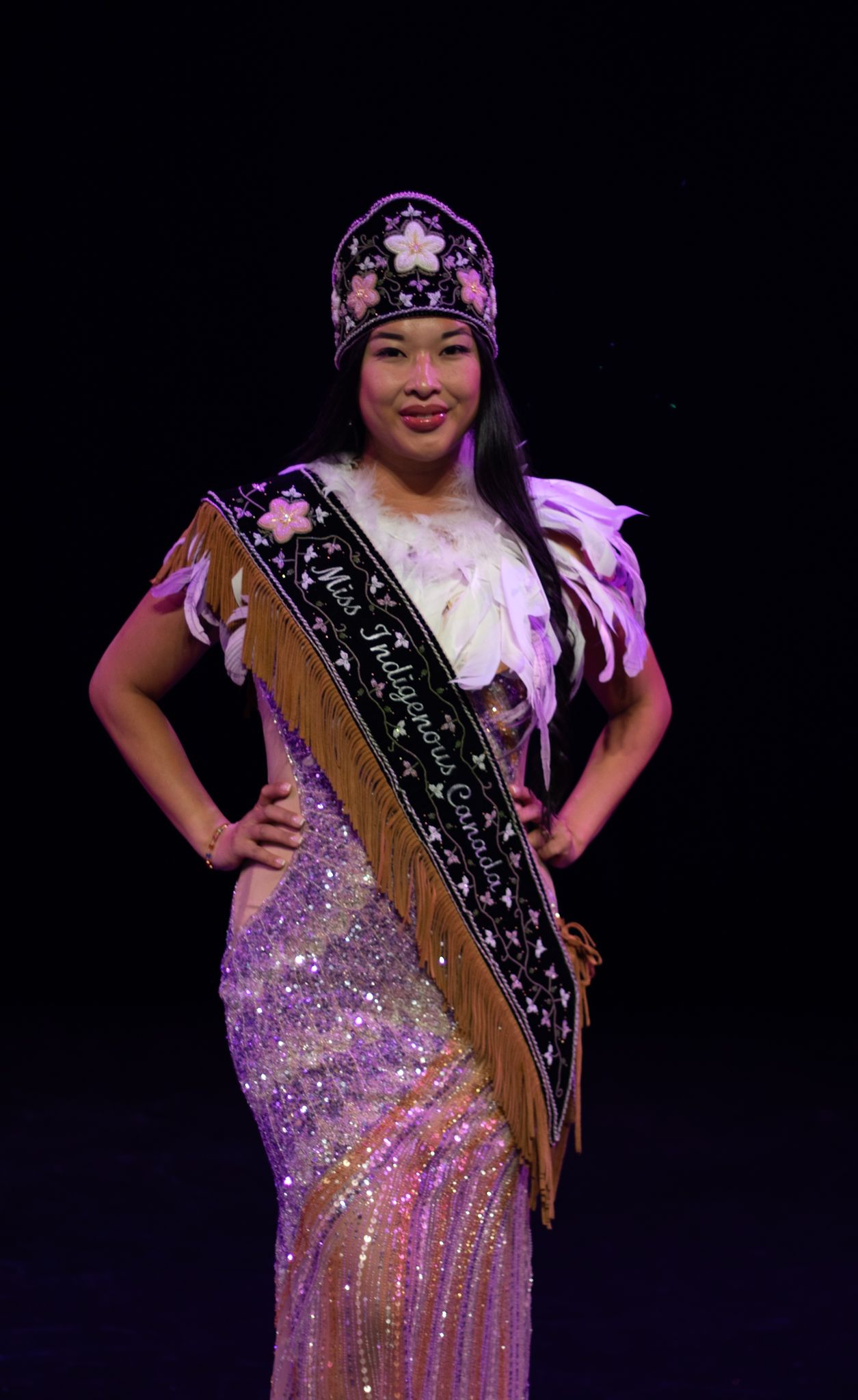 A pageant winner poses in a sparkly gown with sash and beaded crown against dark background.