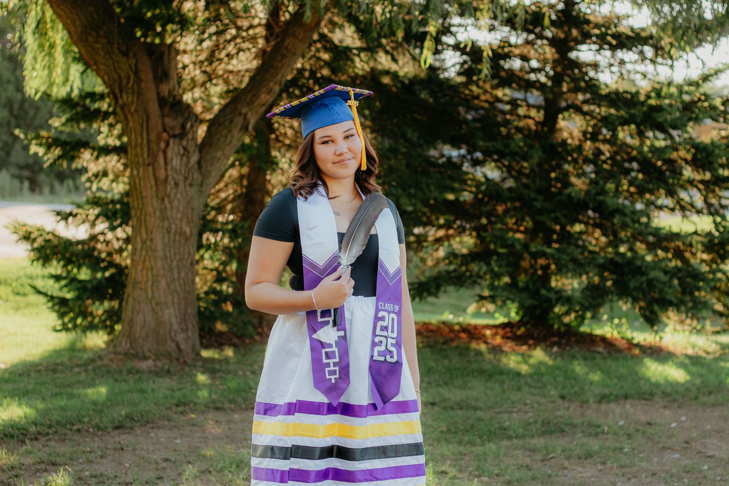 Graduate wearing a varsity jacket and dress poses outdoors near evergreen trees.