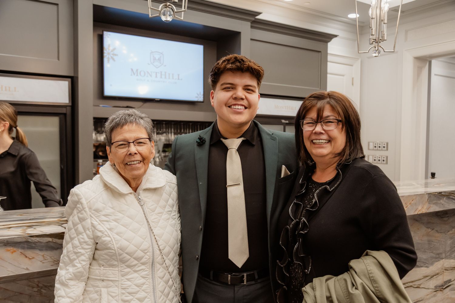 Three people pose together at an elegant venue with mounted screens visible in the background.