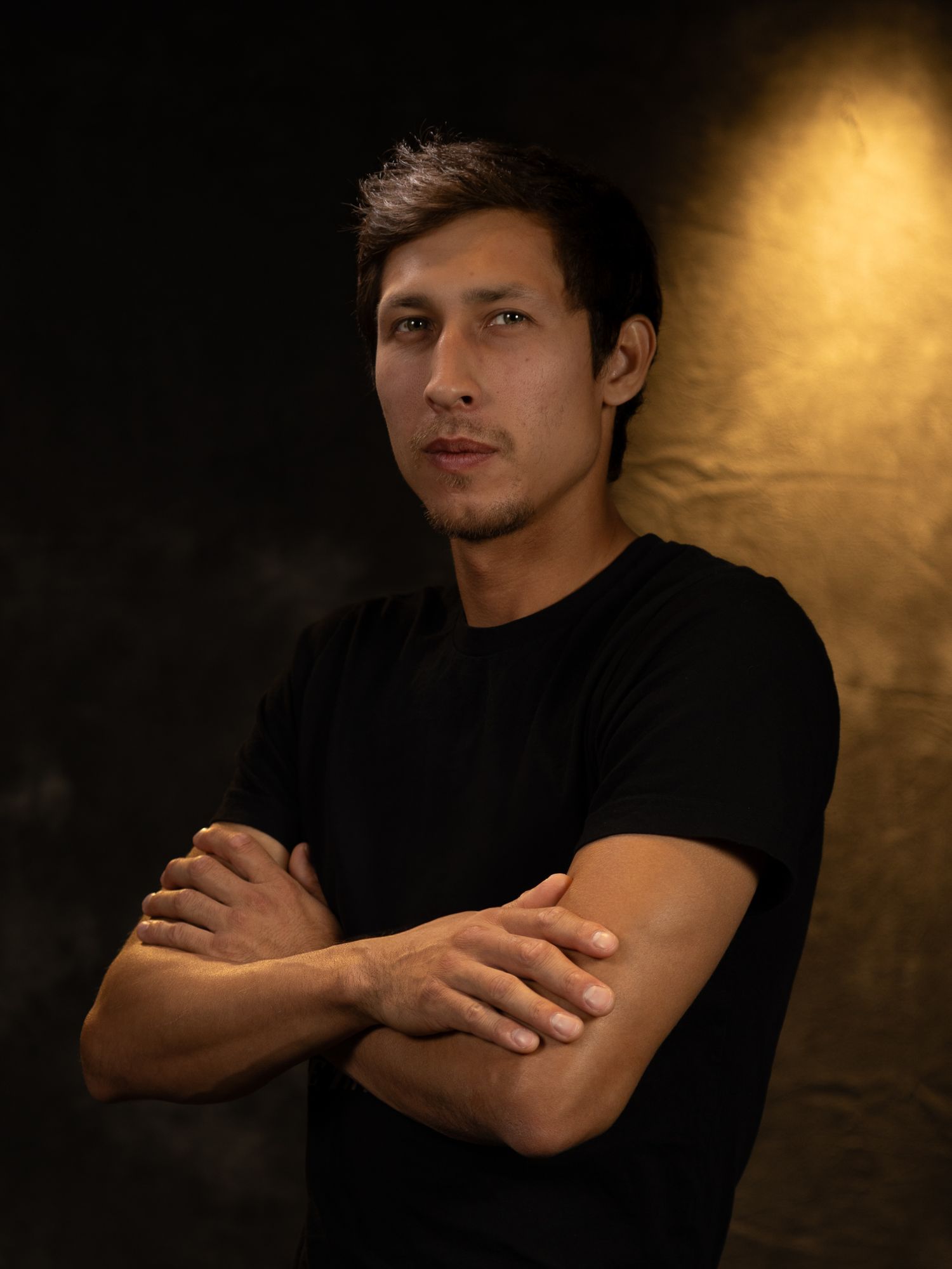 Studio portrait of person in black shirt with arms crossed against dramatic dark background.