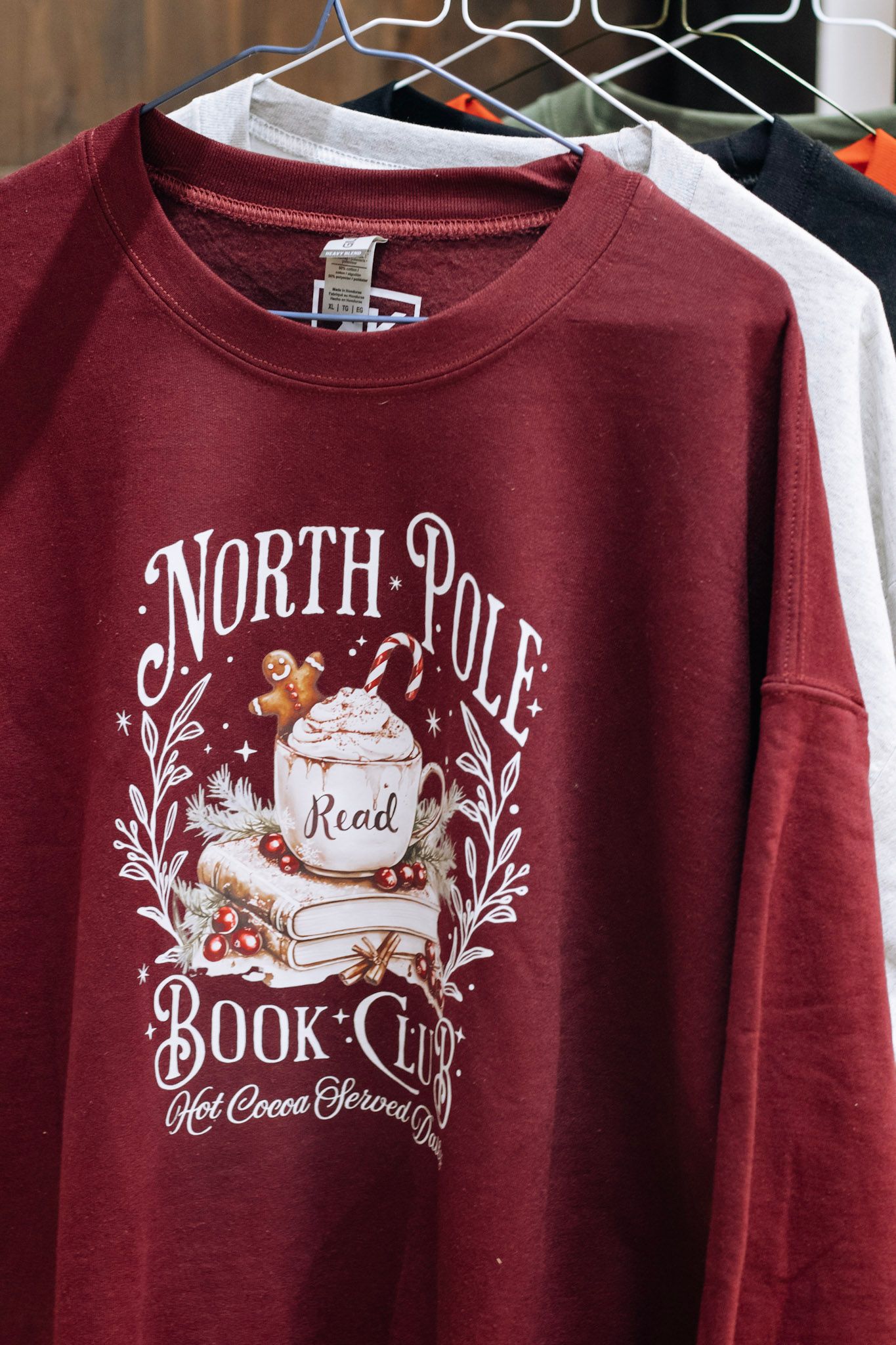 Burgundy holiday sweatshirts with North Pole book club and hot cocoa designs on display.