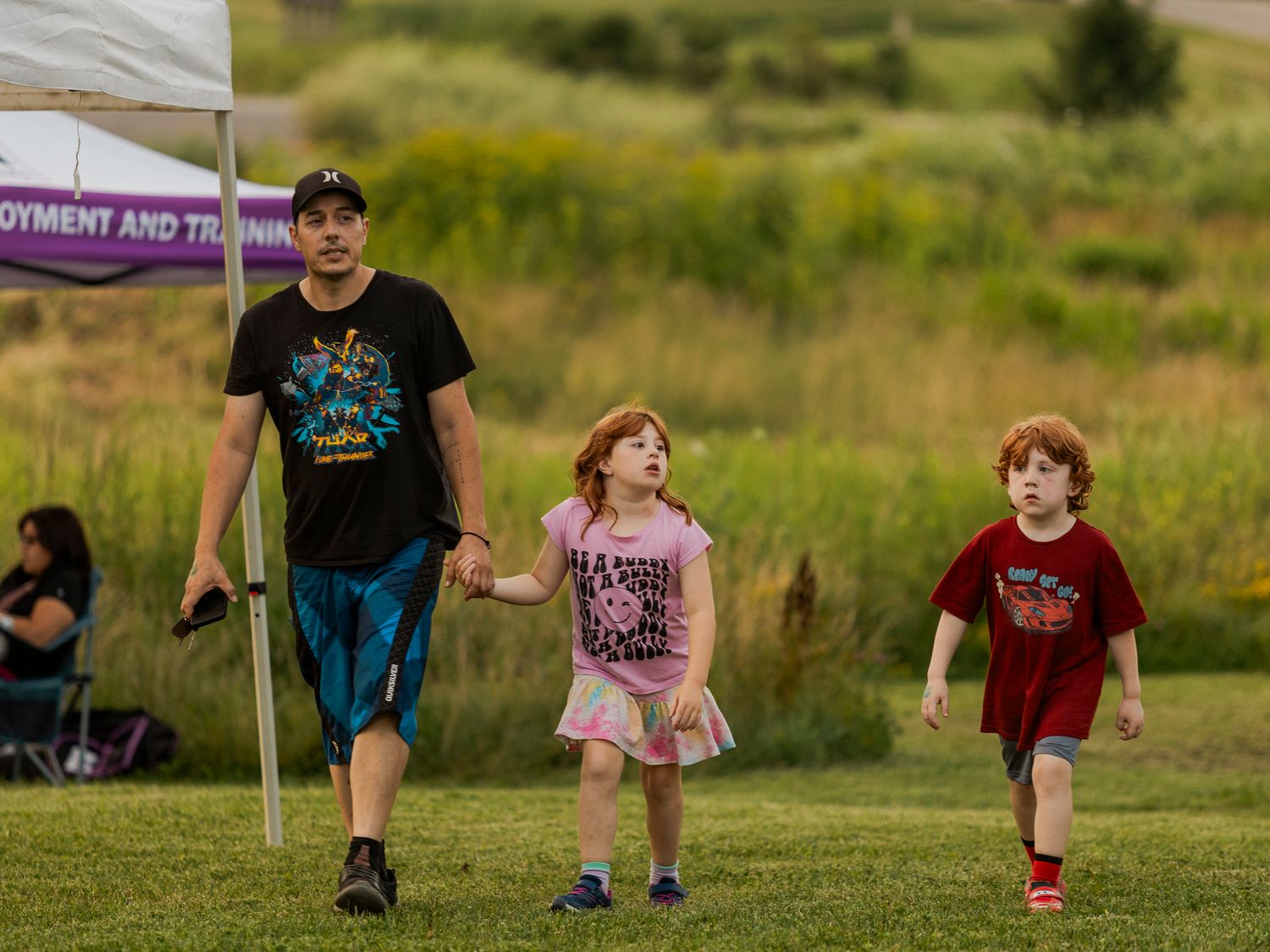 Man walking with two young children on grass near a white tent at an outdoor community event.