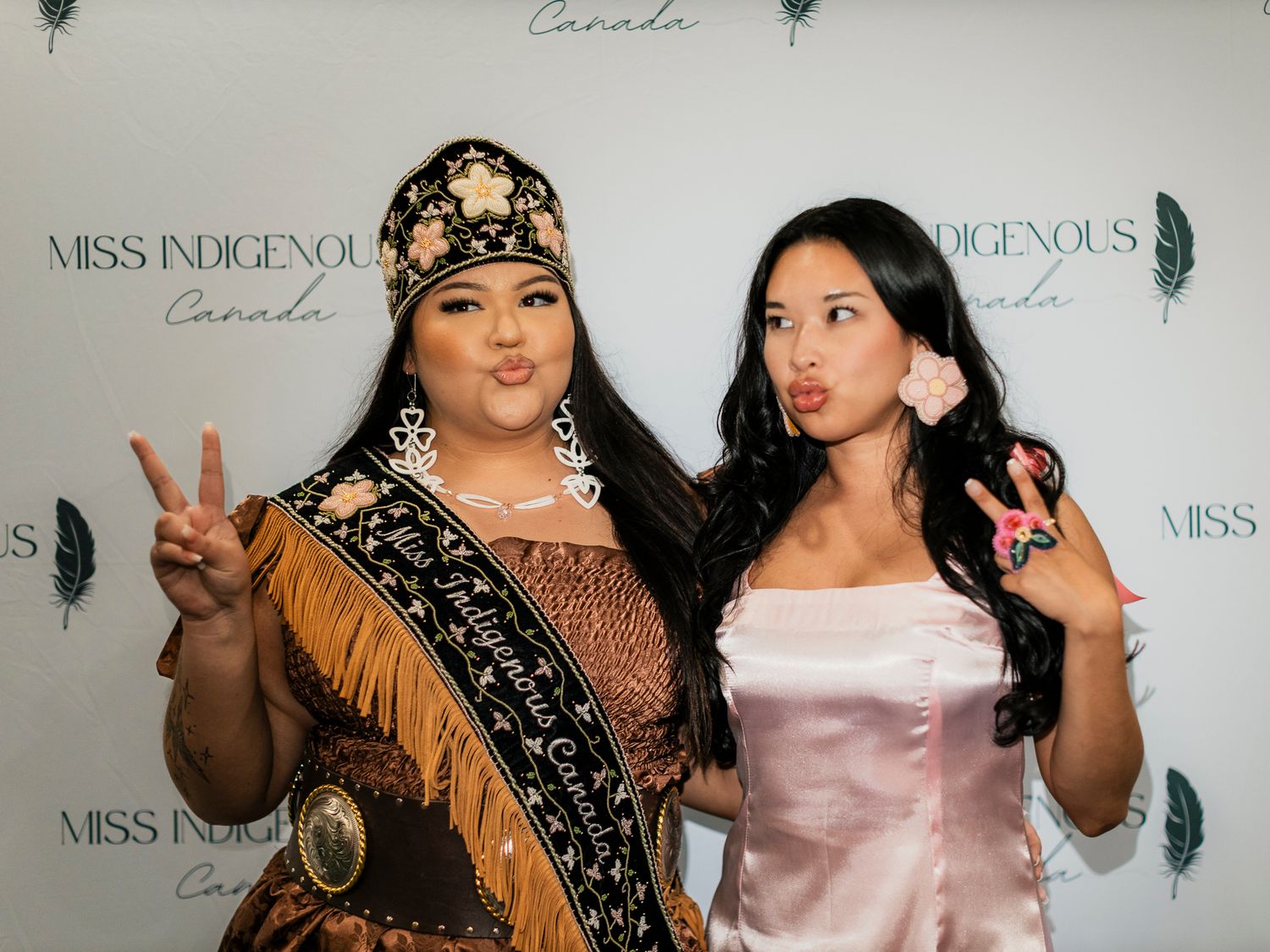 Two women pose at Miss Indigenous Canada event, one wearing a sash and traditional regalia, both making peace signs.
