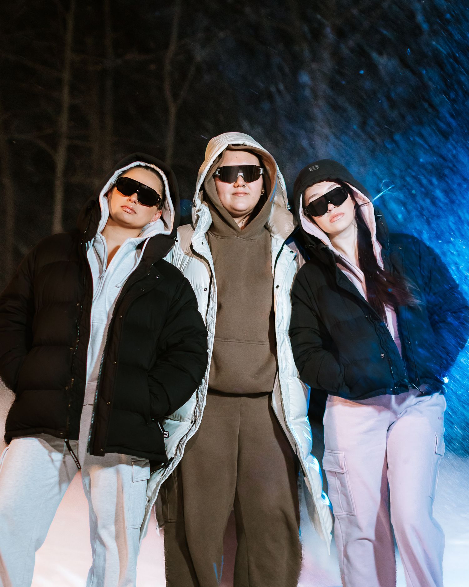 Three people in hooded puffer jackets and sunglasses posing in snowy night setting.