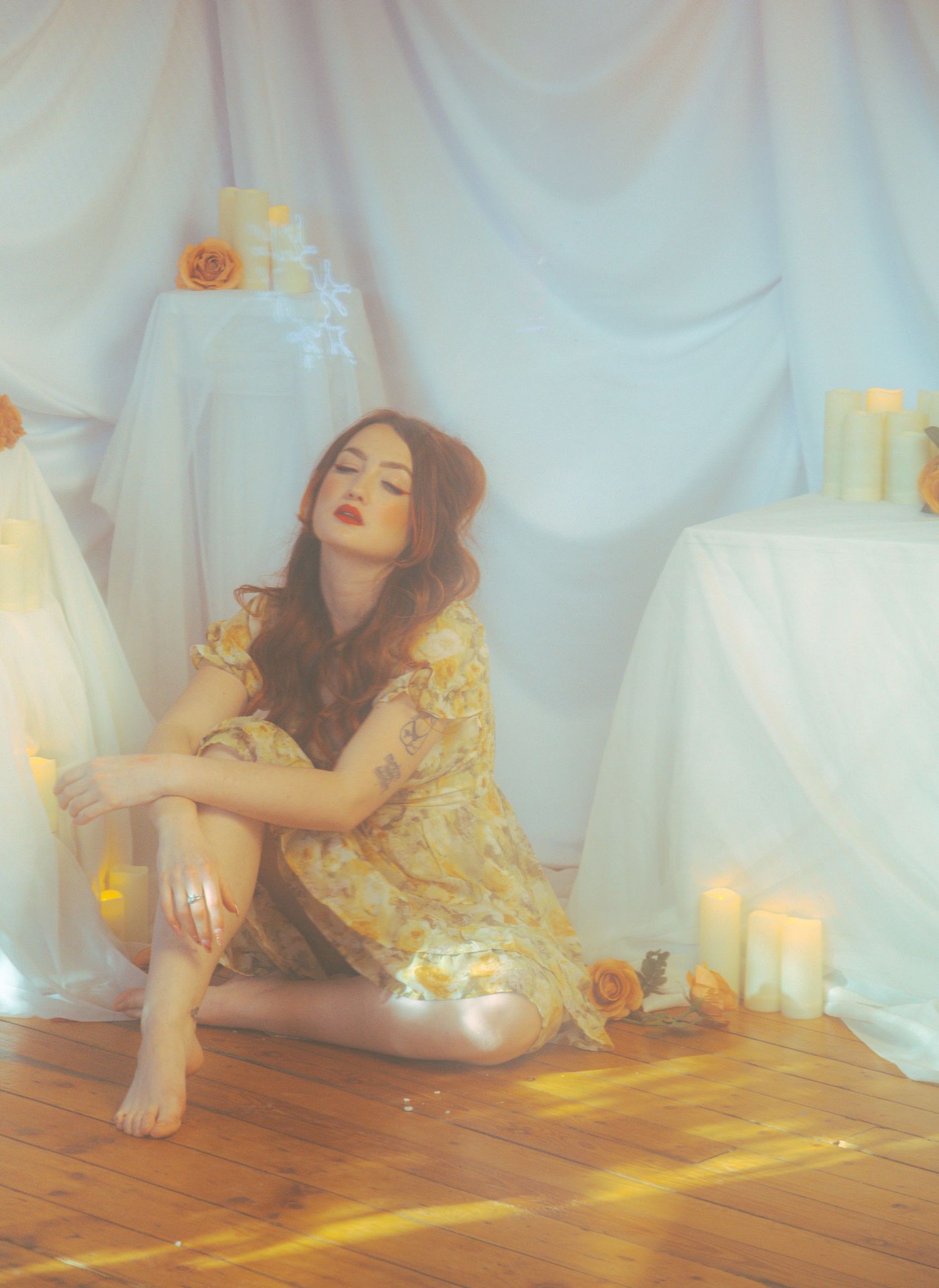 A woman in a floral yellow dress sits gracefully on a wooden floor surrounded by candles and white draping.