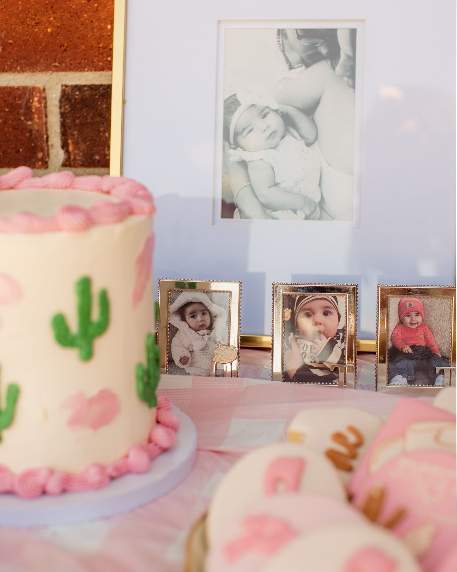 Pink birthday cake with cactus decorations displayed with baby photo frames on a dessert table.