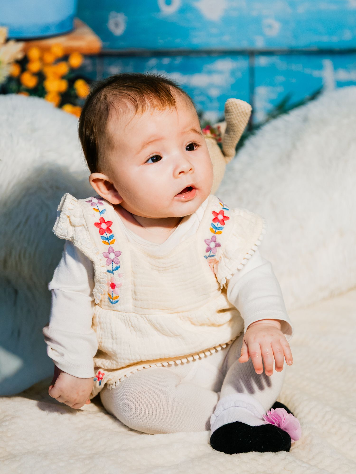 Adorable baby girl in cream floral embroidered outfit sitting on white fluffy blanket with blue backdrop.