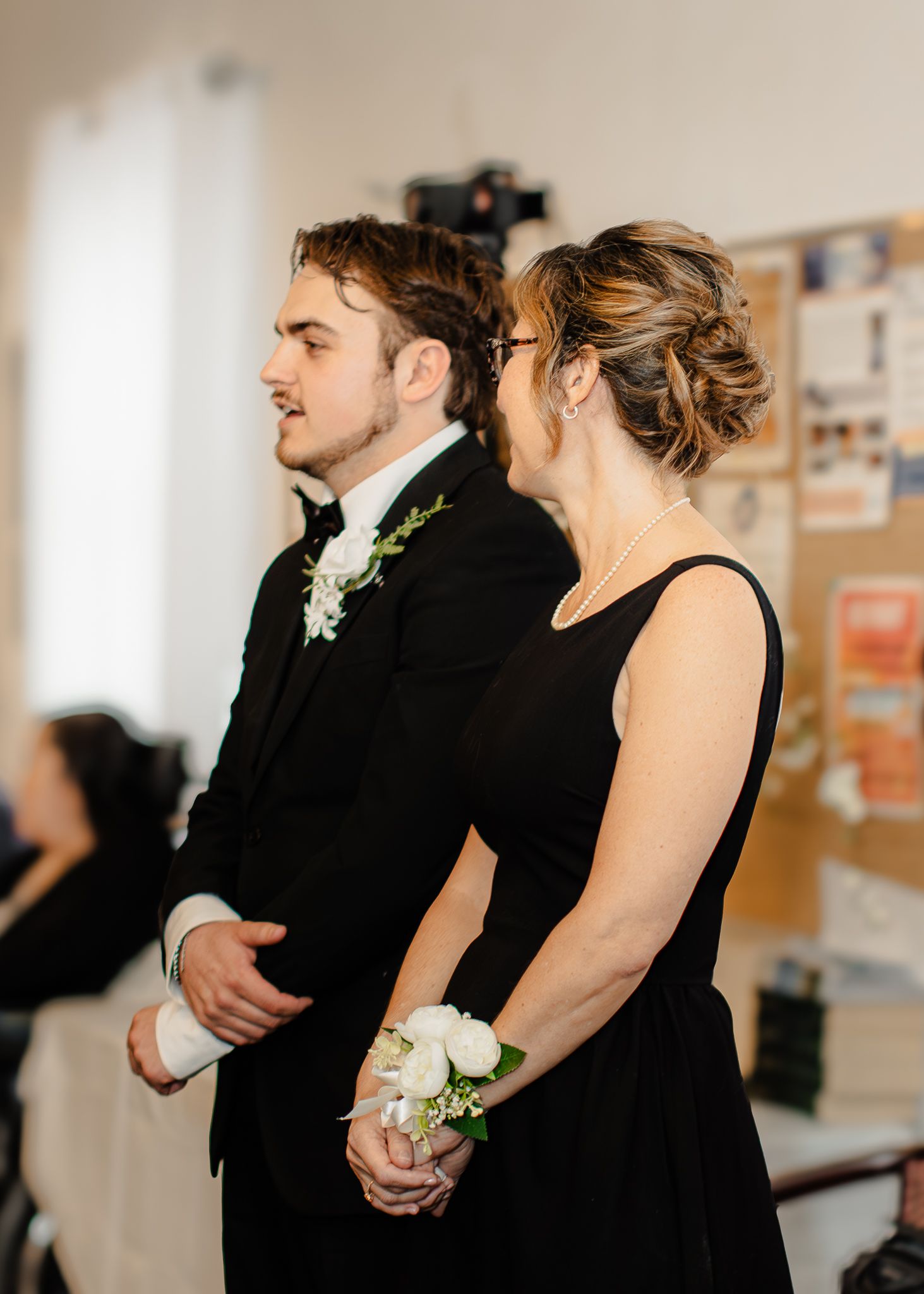 Groom in black tuxedo escorts woman in black dress with white wrist corsage at wedding ceremony.