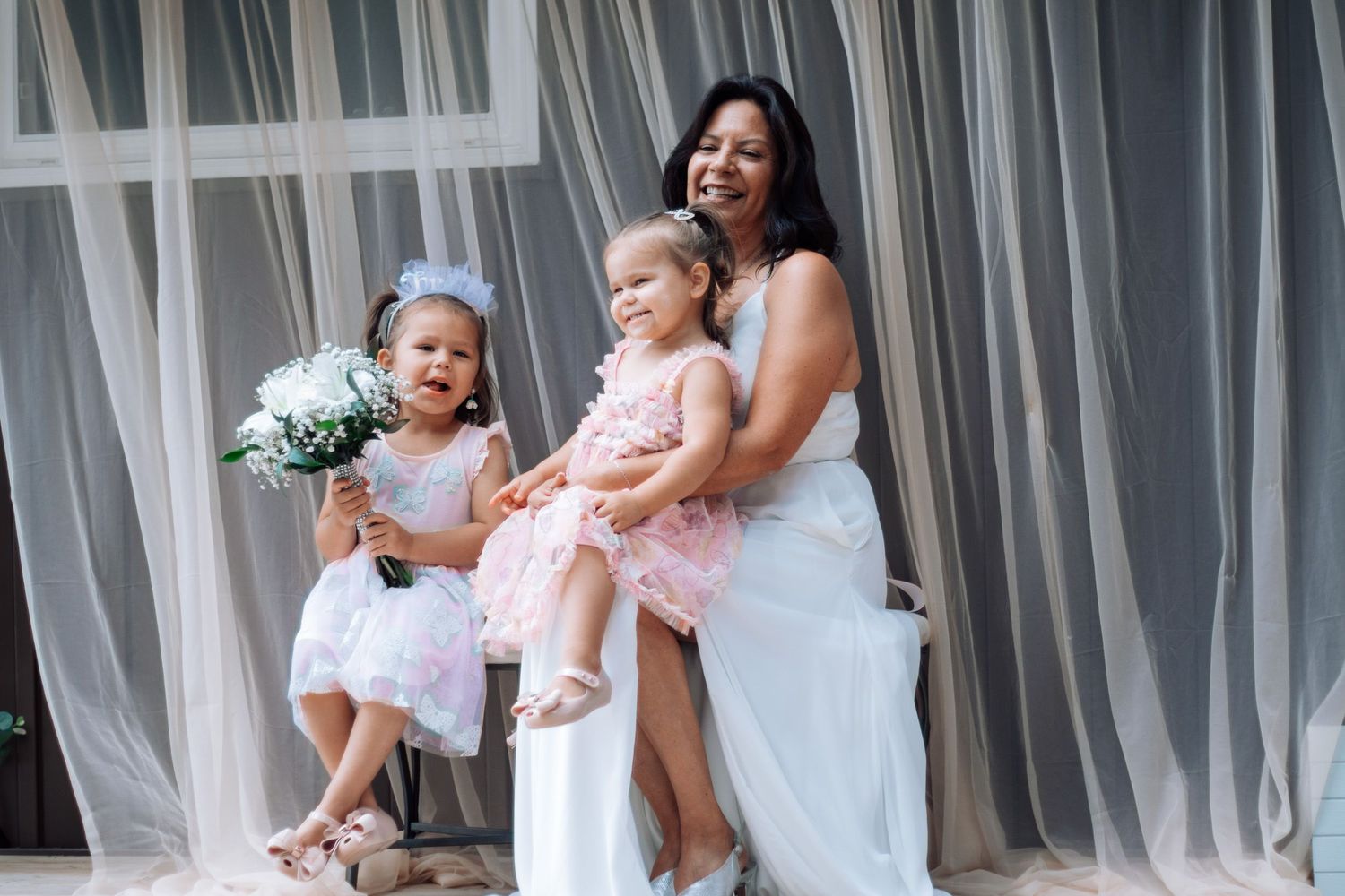Bride in white gown seated with two young flower girls in pink dresses holding white bouquet.