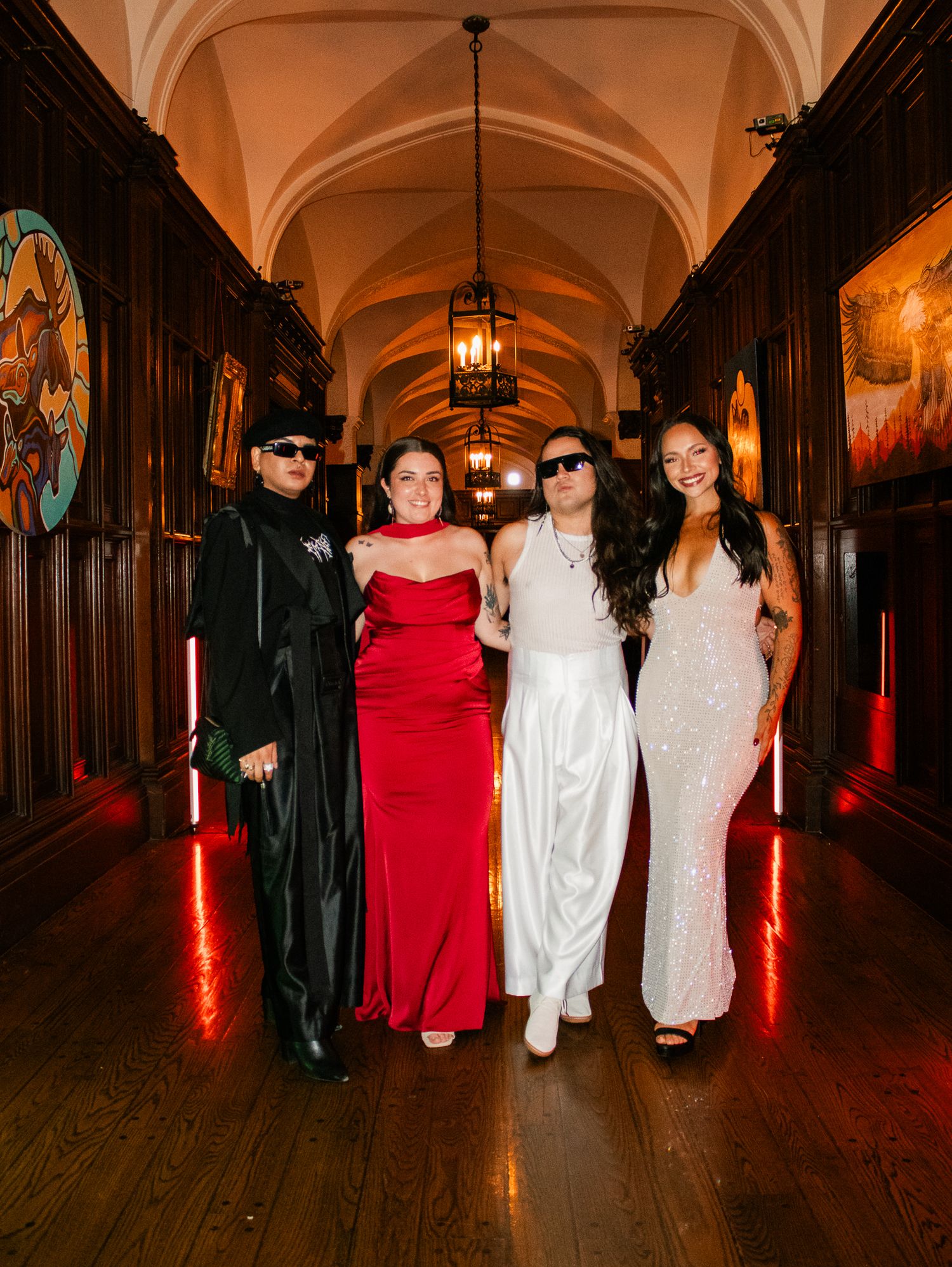 Four friends in formal gowns posing in a grand castle hallway with arched ceilings and warm lighting.