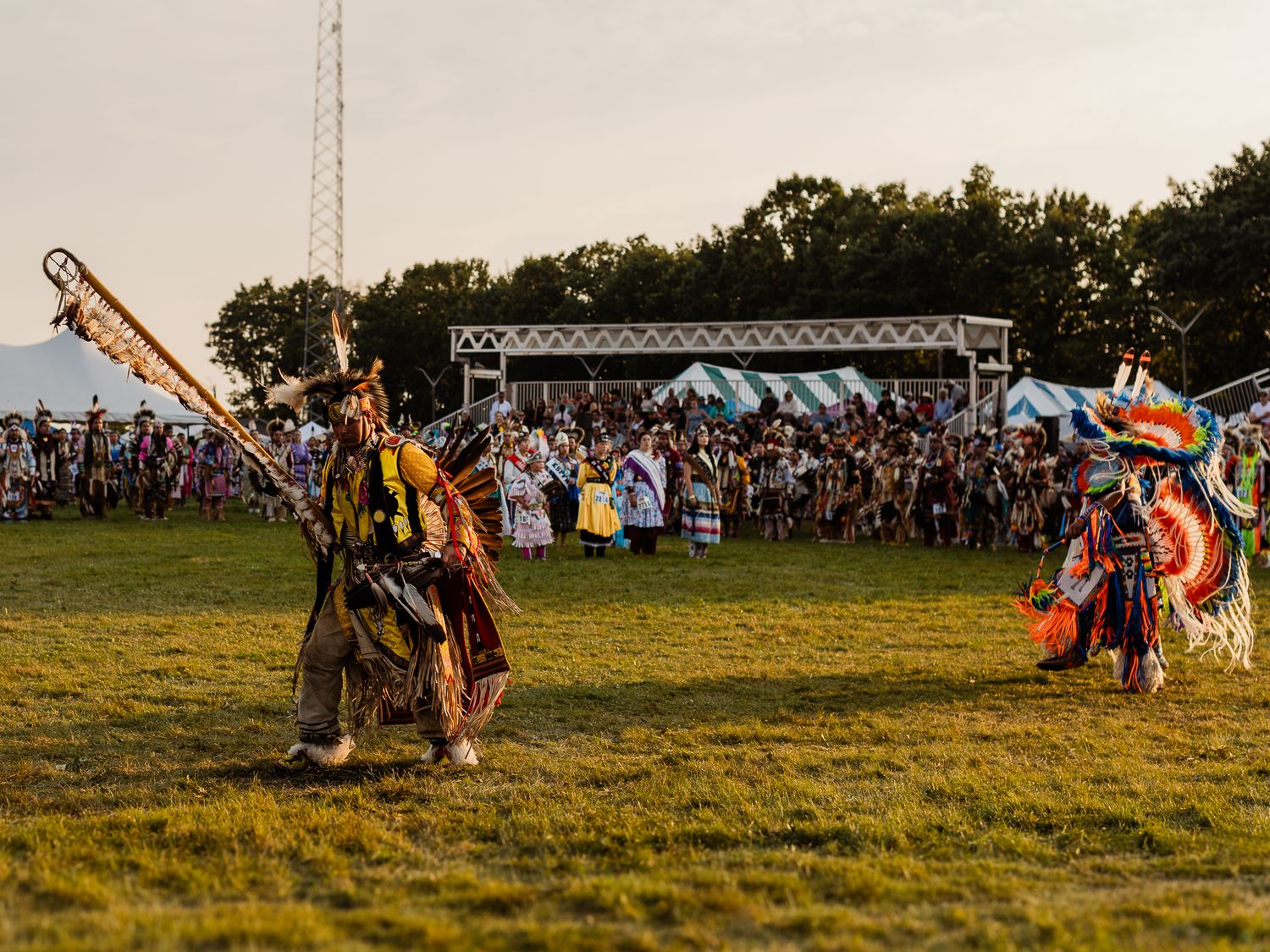Native American dancers in colorful traditional regalia perform at an outdoor powwow ceremony at dusk.
