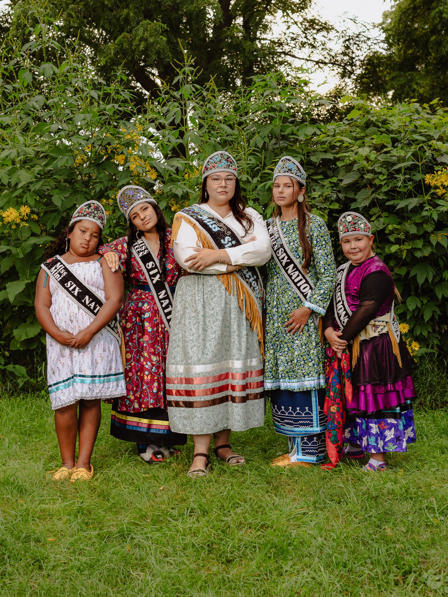 Five women in traditional Native American regalia with sashes pose together outdoors among green foliage.