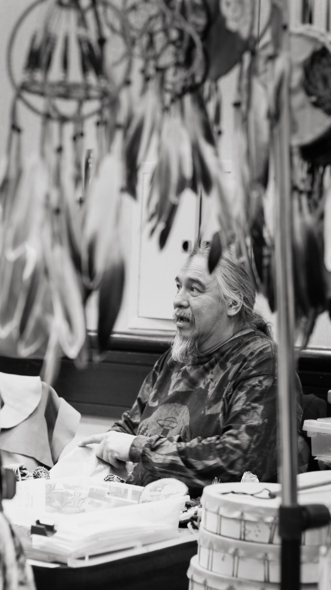 Black and white photo of an older Native American man sitting among handmade dreamcatchers with feathers at a craft market.