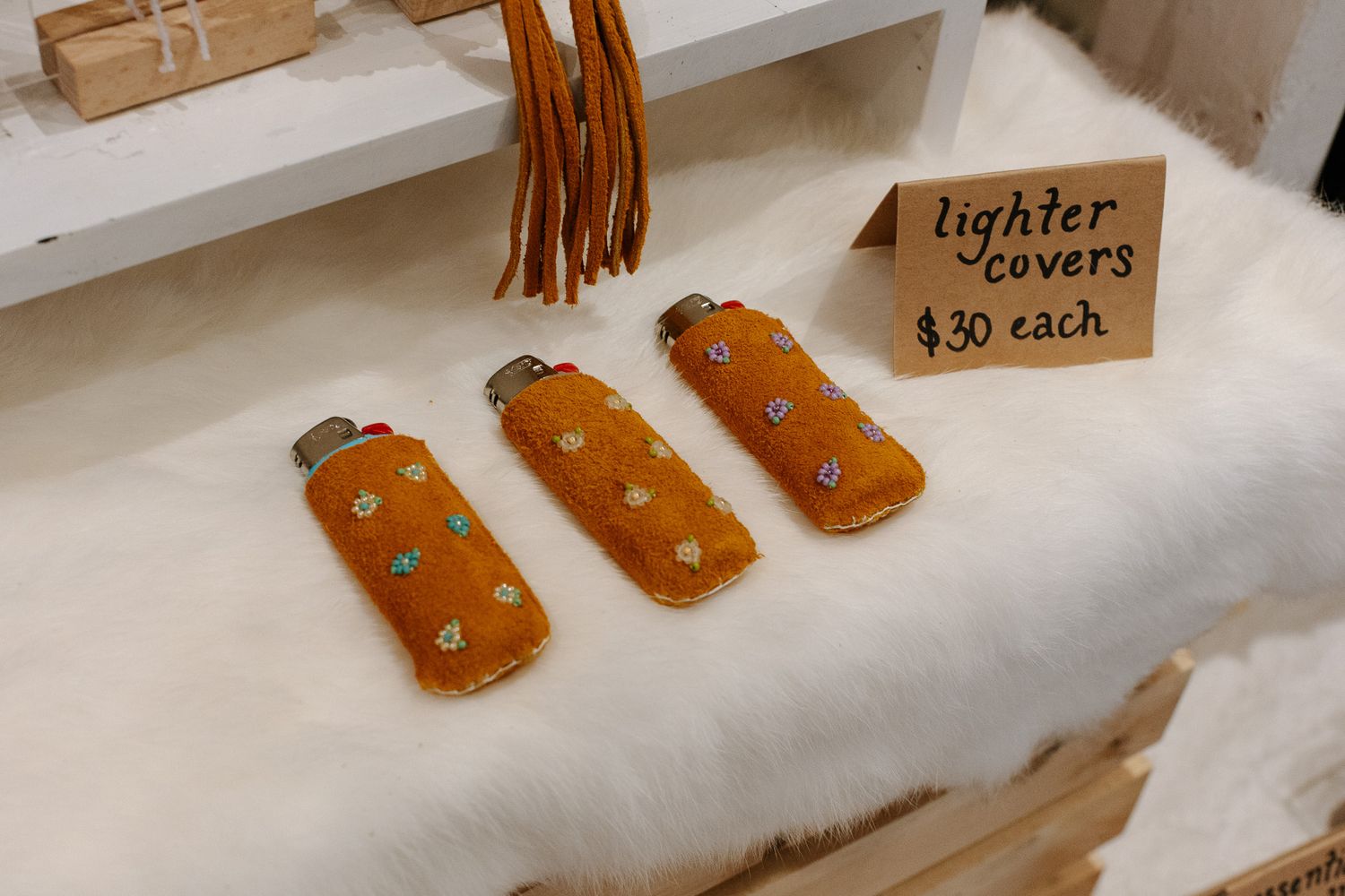 Three orange leather lighter covers with turquoise beading displayed on white fur, priced at $30 each.
