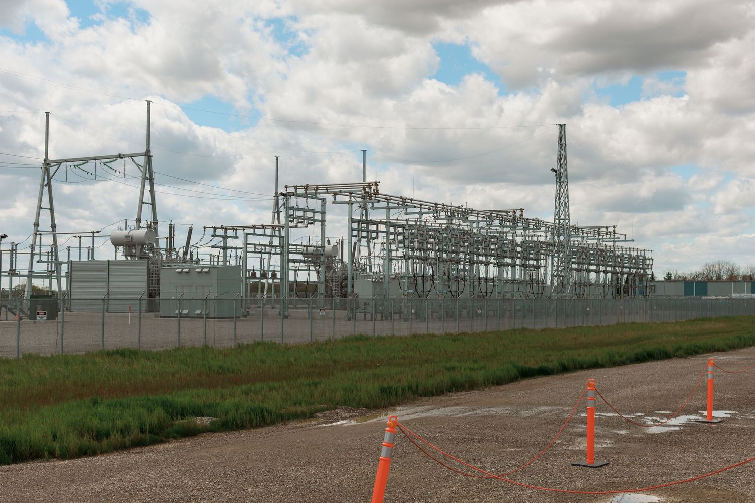 Large electrical substation with steel framework and power lines behind security fence under cloudy sky.