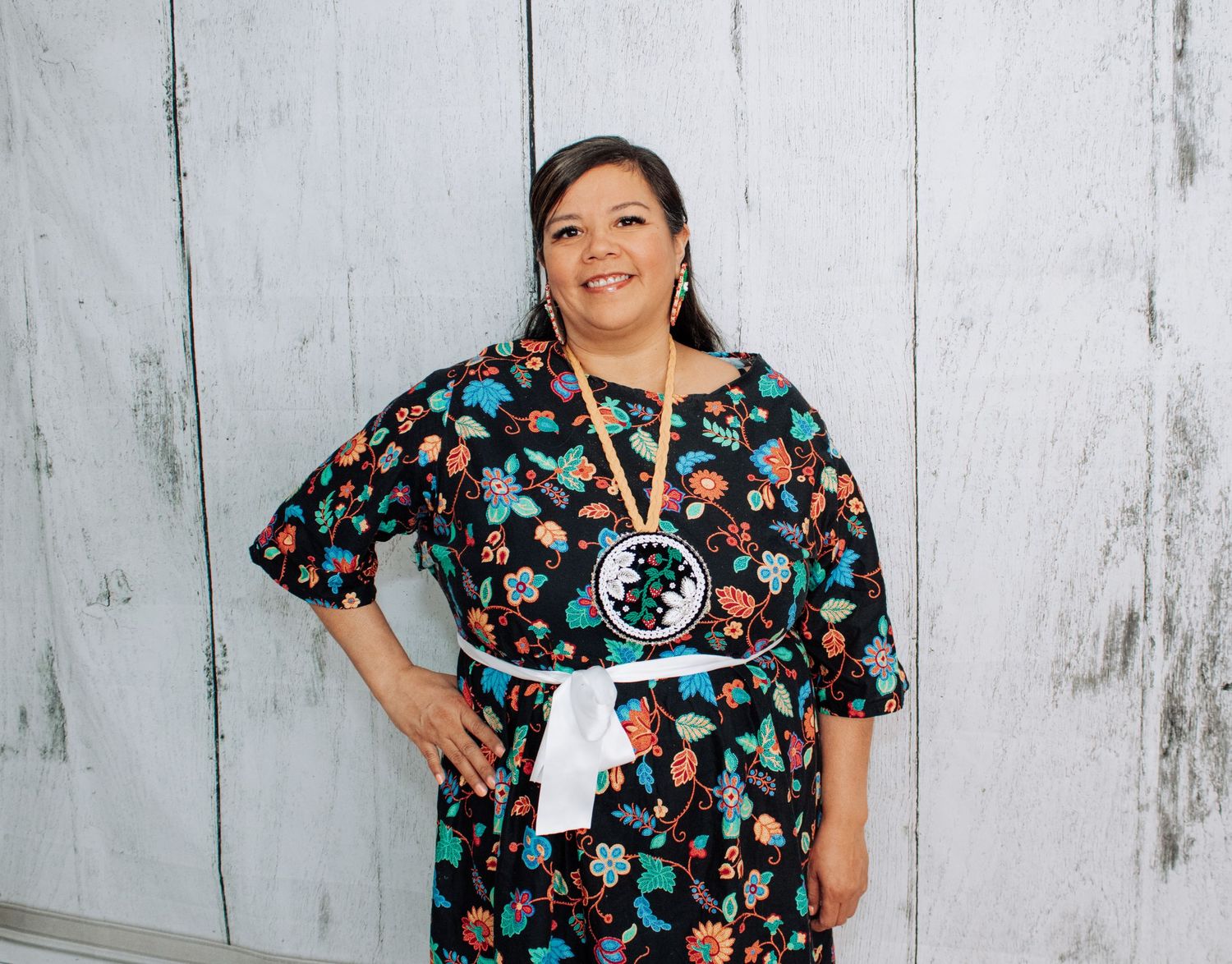 A person wearing a floral patterned dress with traditional Native American accessories poses against a wall.