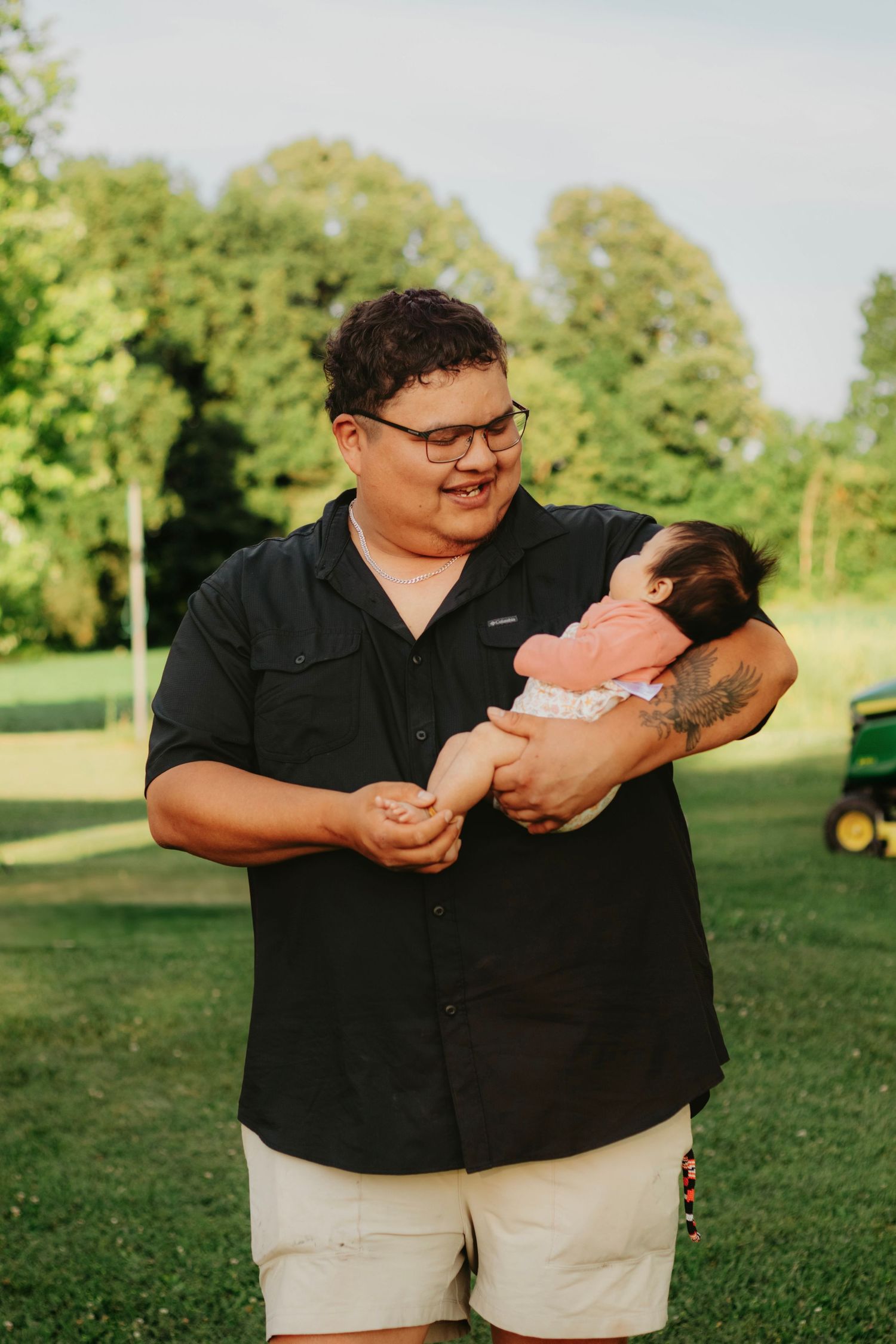 Person in black shirt holding a newborn baby outdoors on green grass with trees in background.