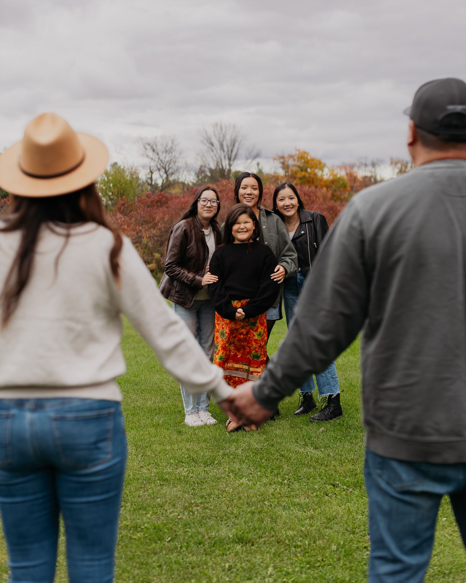 Family gathering outdoors on green grass in autumn, holding hands in a circle with colorful fall foliage in background.