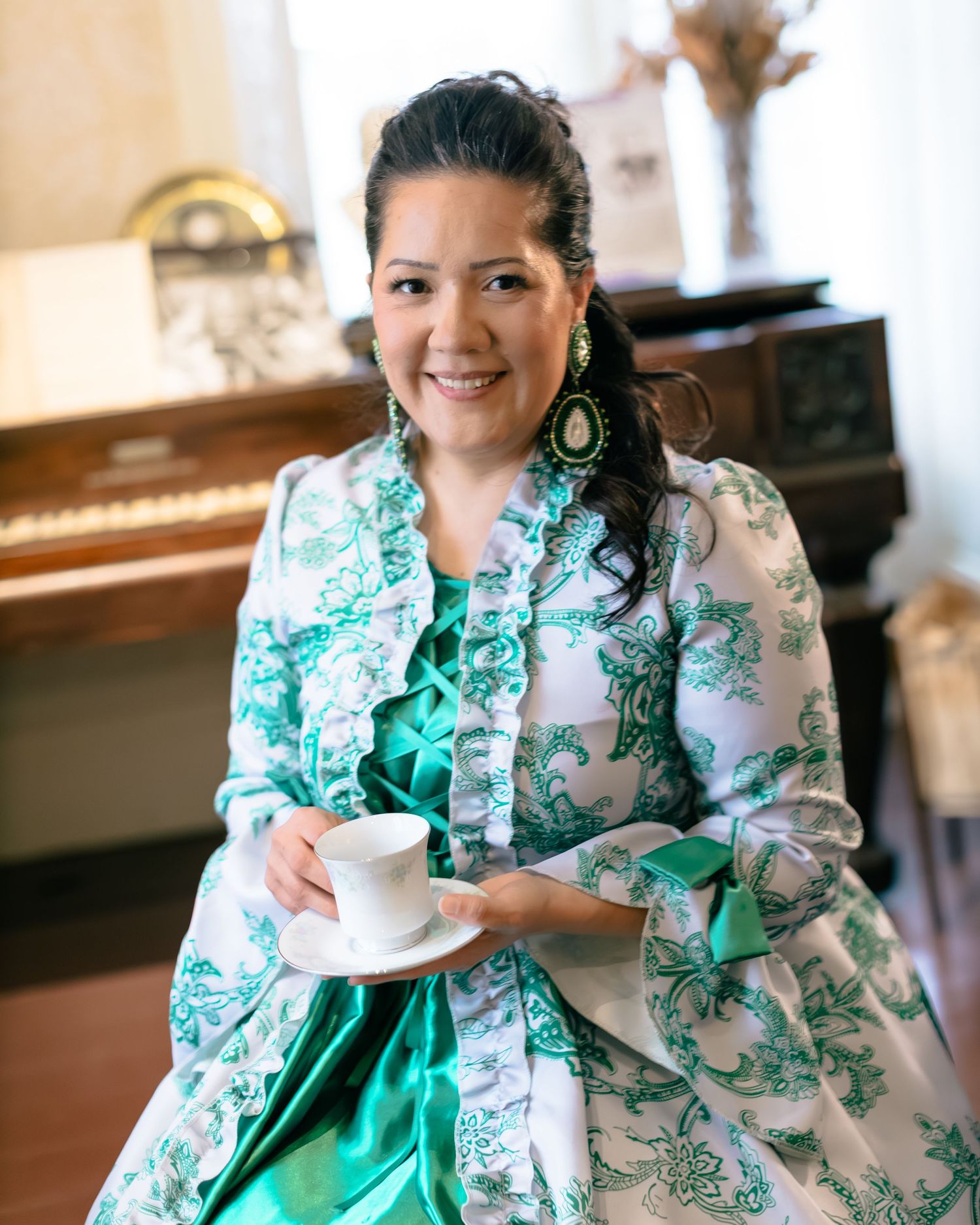 Woman in elegant green and white floral dress holding a teacup near a vintage piano.
