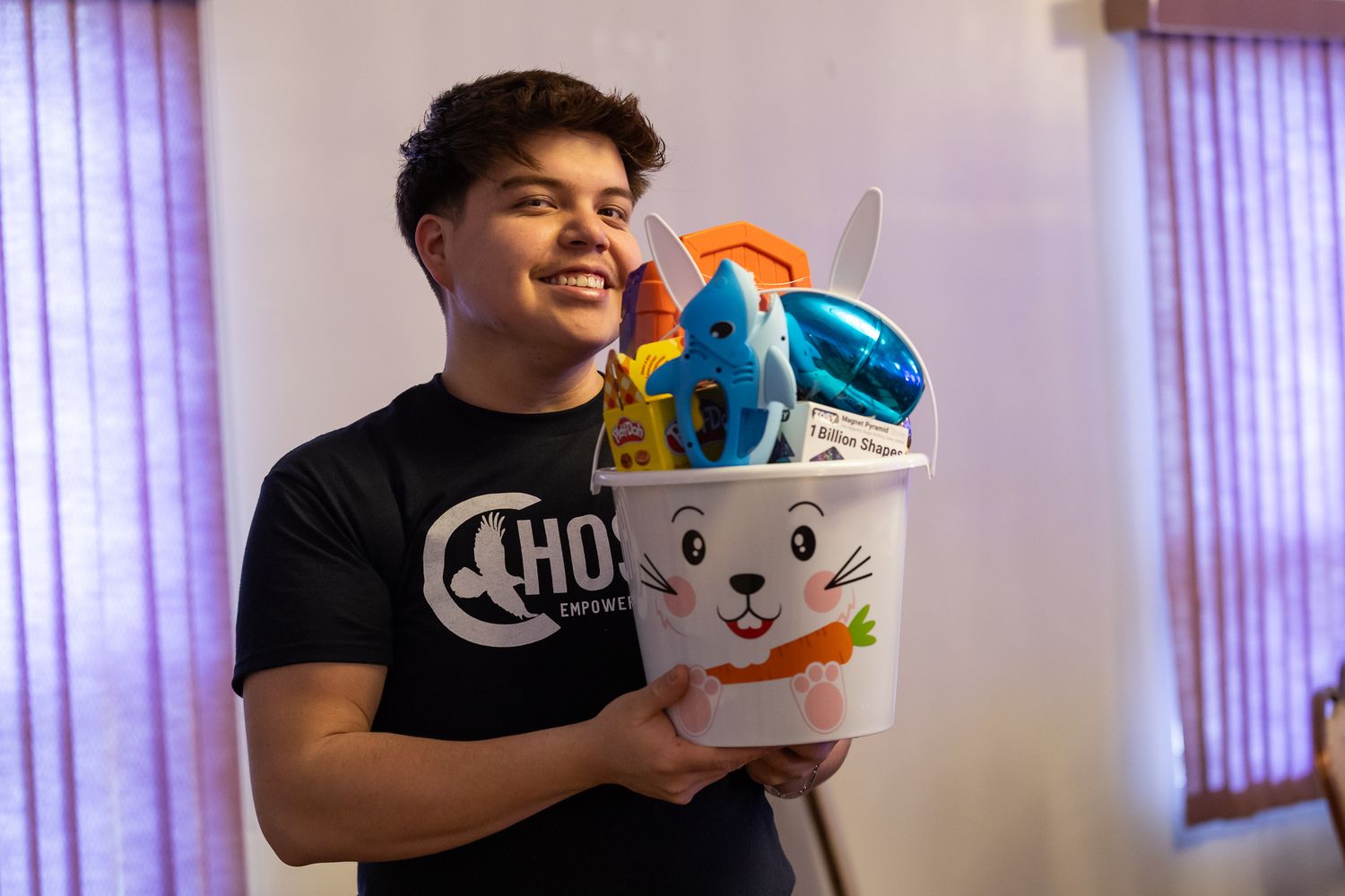 Smiling teen holding a white Easter bunny basket filled with colorful gifts and toys.