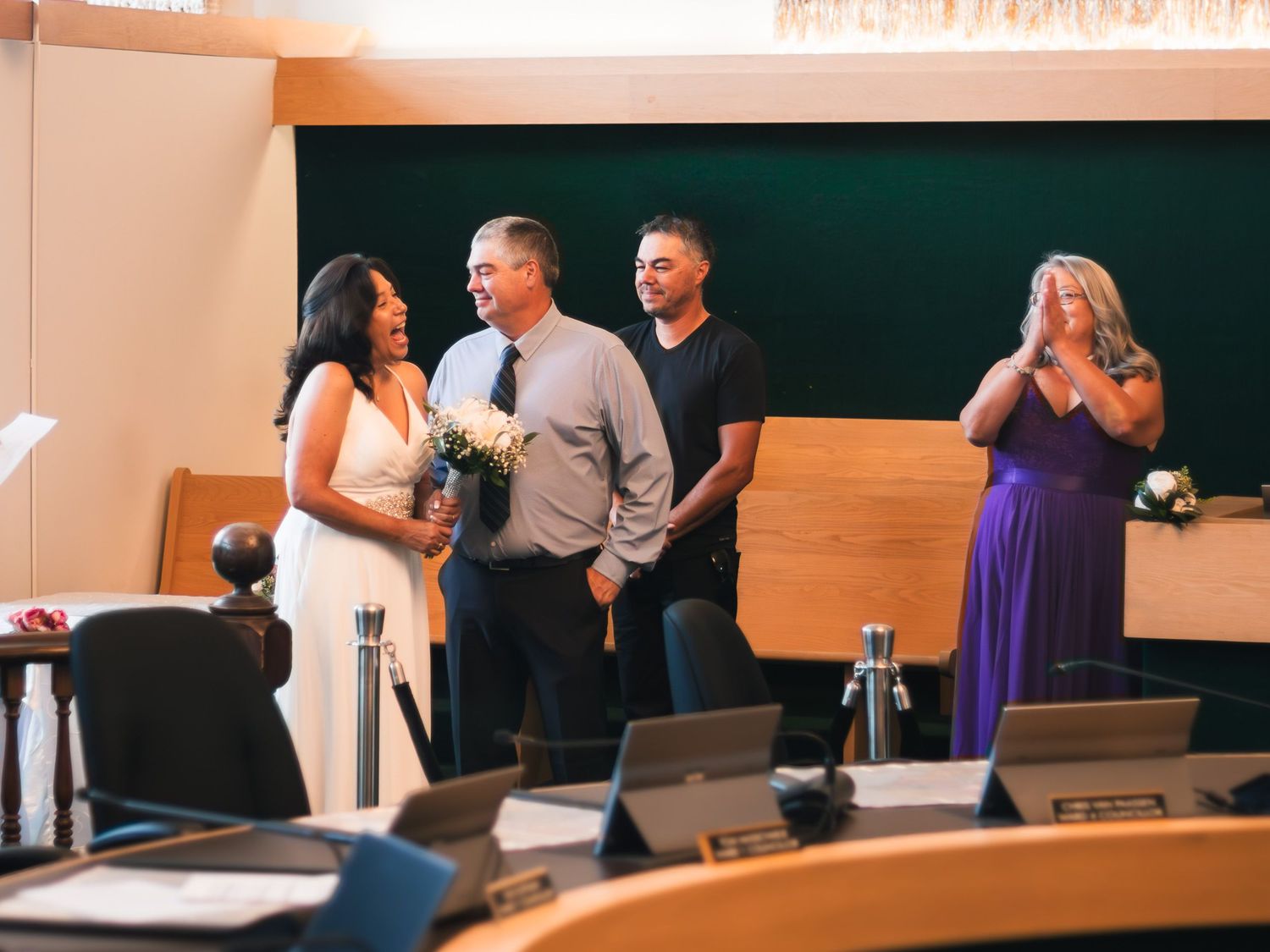 Wedding ceremony with bride holding bouquet, groom, officiant, and guests clapping in modern venue.