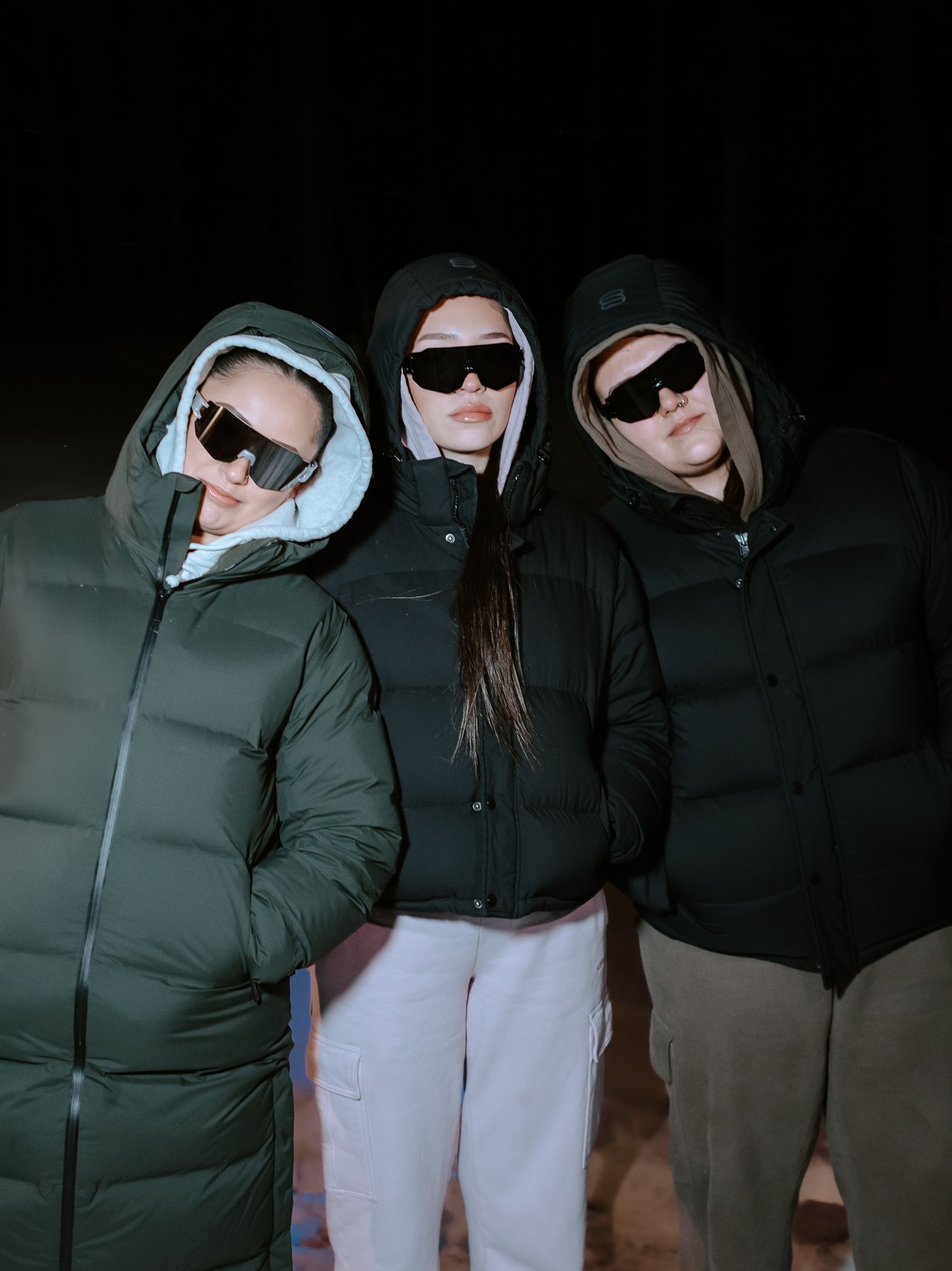 Three women wearing puffer jackets, hoods, and sunglasses pose together against a dark night sky.