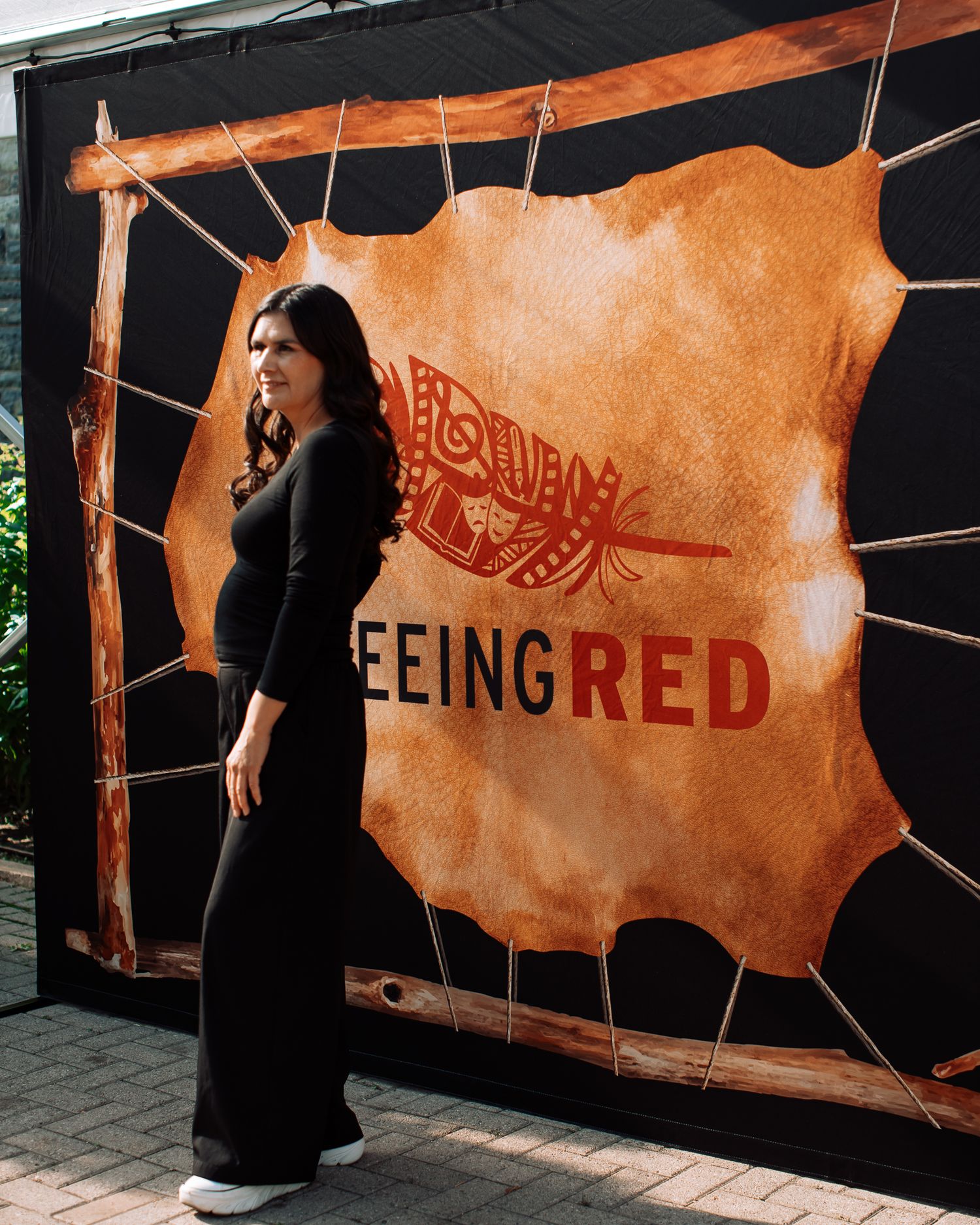 Woman in black outfit poses before Seeing Red Indigenous art exhibition backdrop with leather hide display.