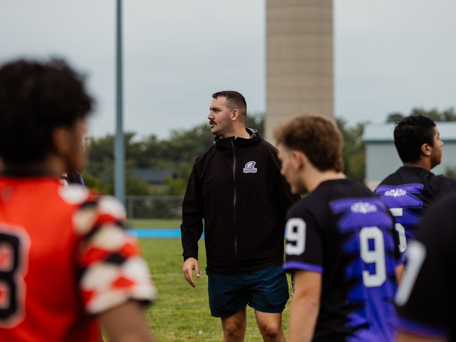 Coach in black jacket observing players in orange and purple jerseys during an outdoor rugby practice session.