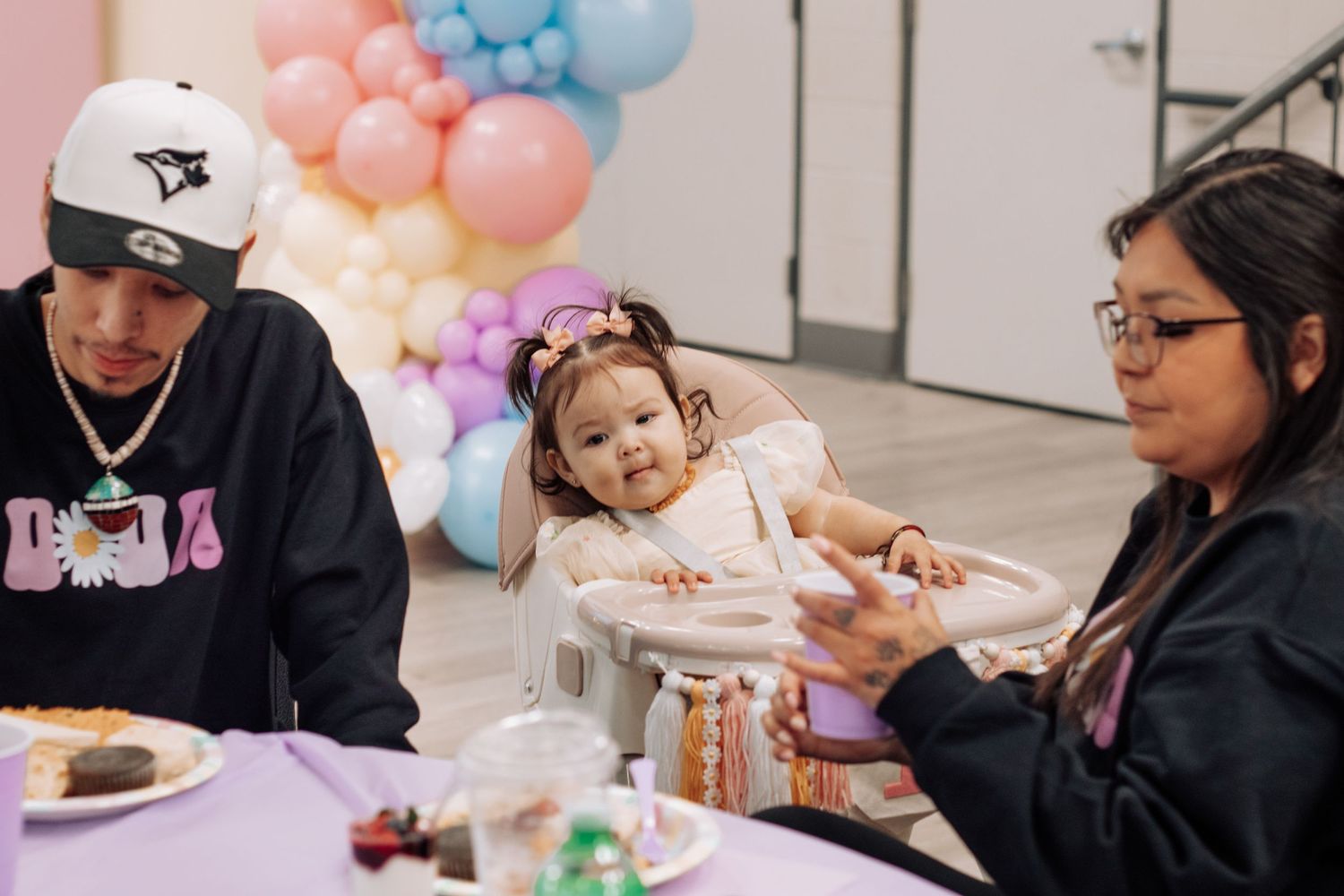 A baby girl in a high chair celebrates her first birthday with family at a pastel balloon-decorated party table.