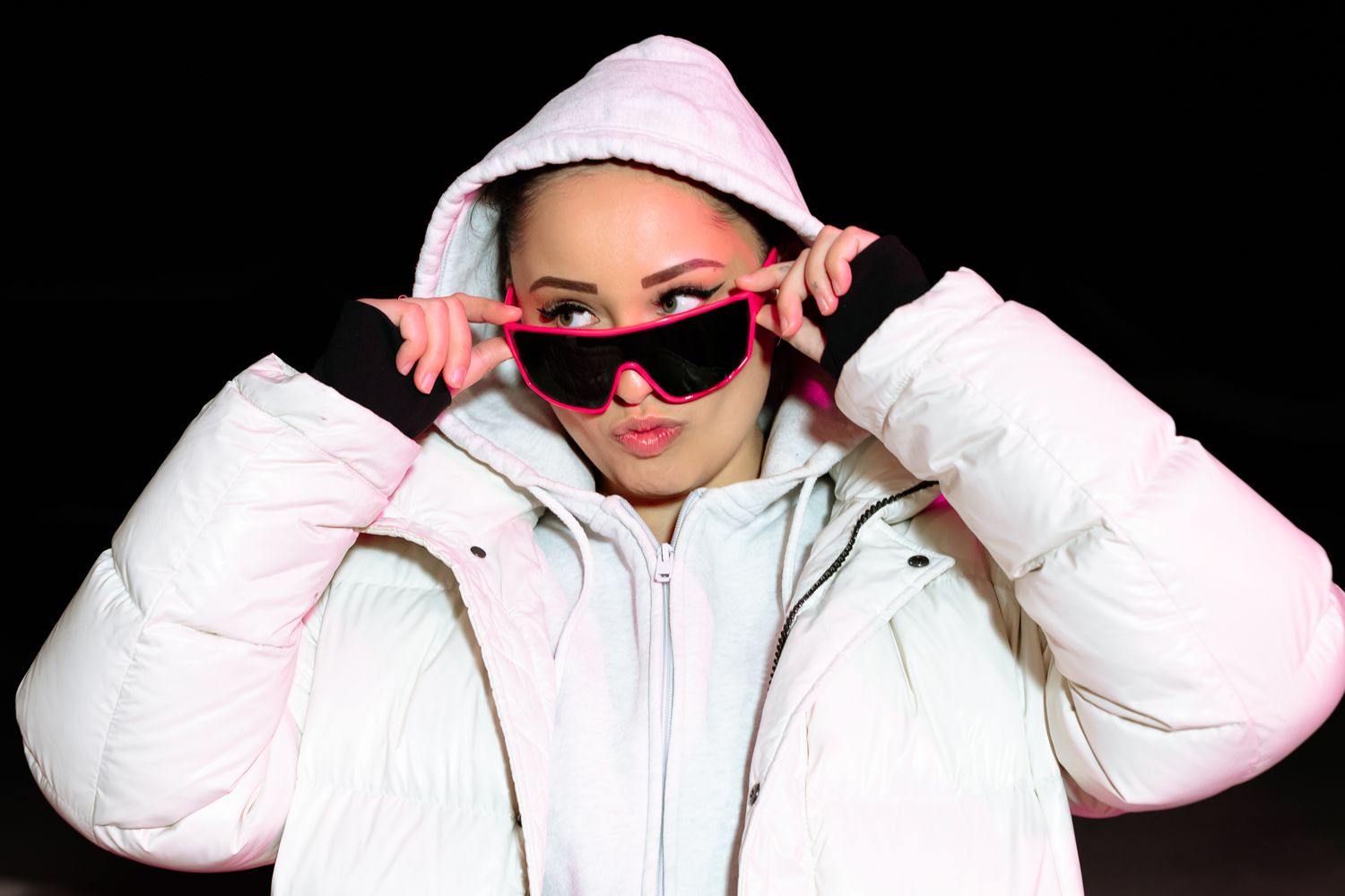 Woman in white hooded puffer jacket adjusting pink sunglasses against dark background.