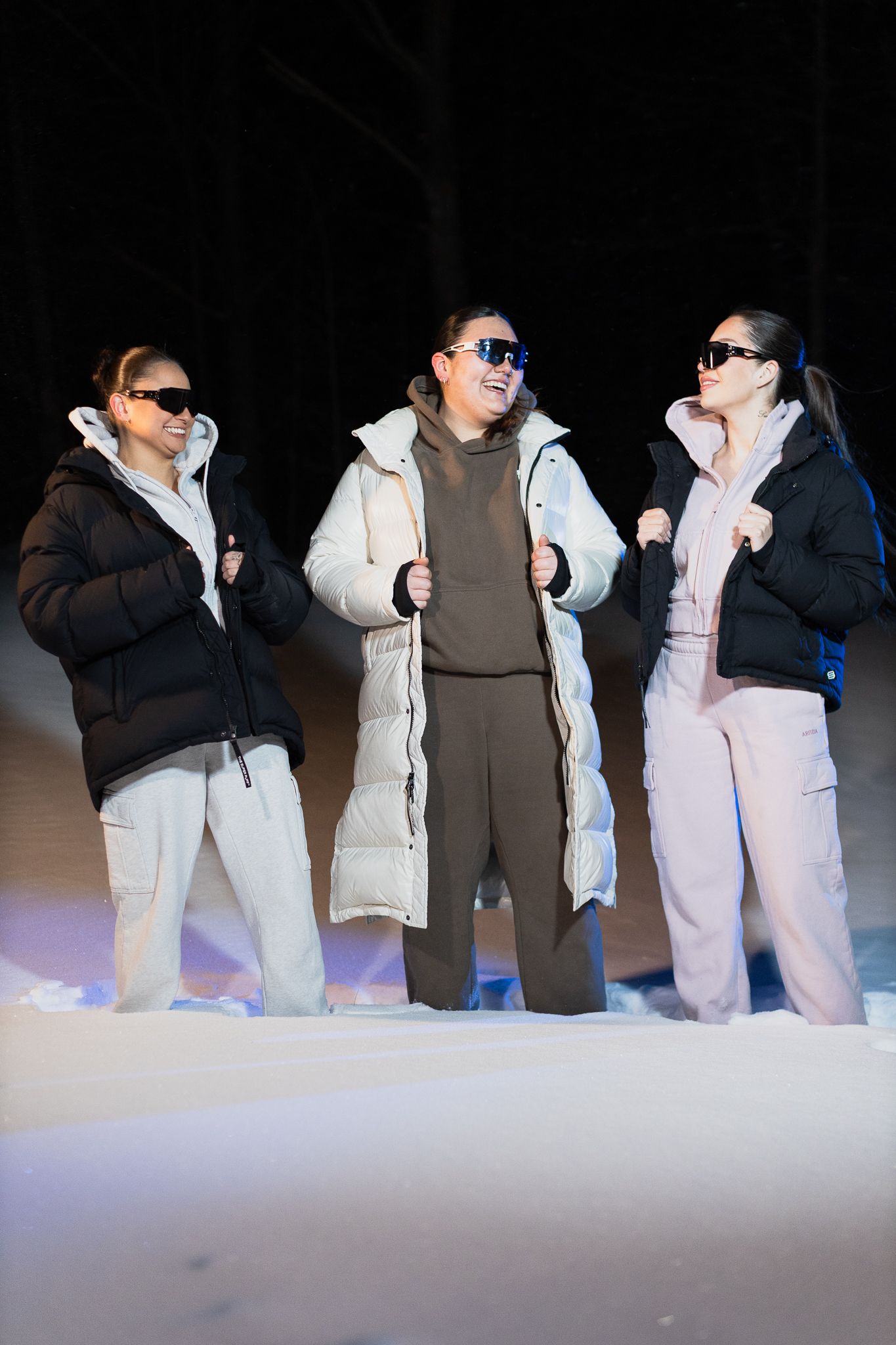 Three smiling women wearing puffer coats and sunglasses stand together in snowy night setting.