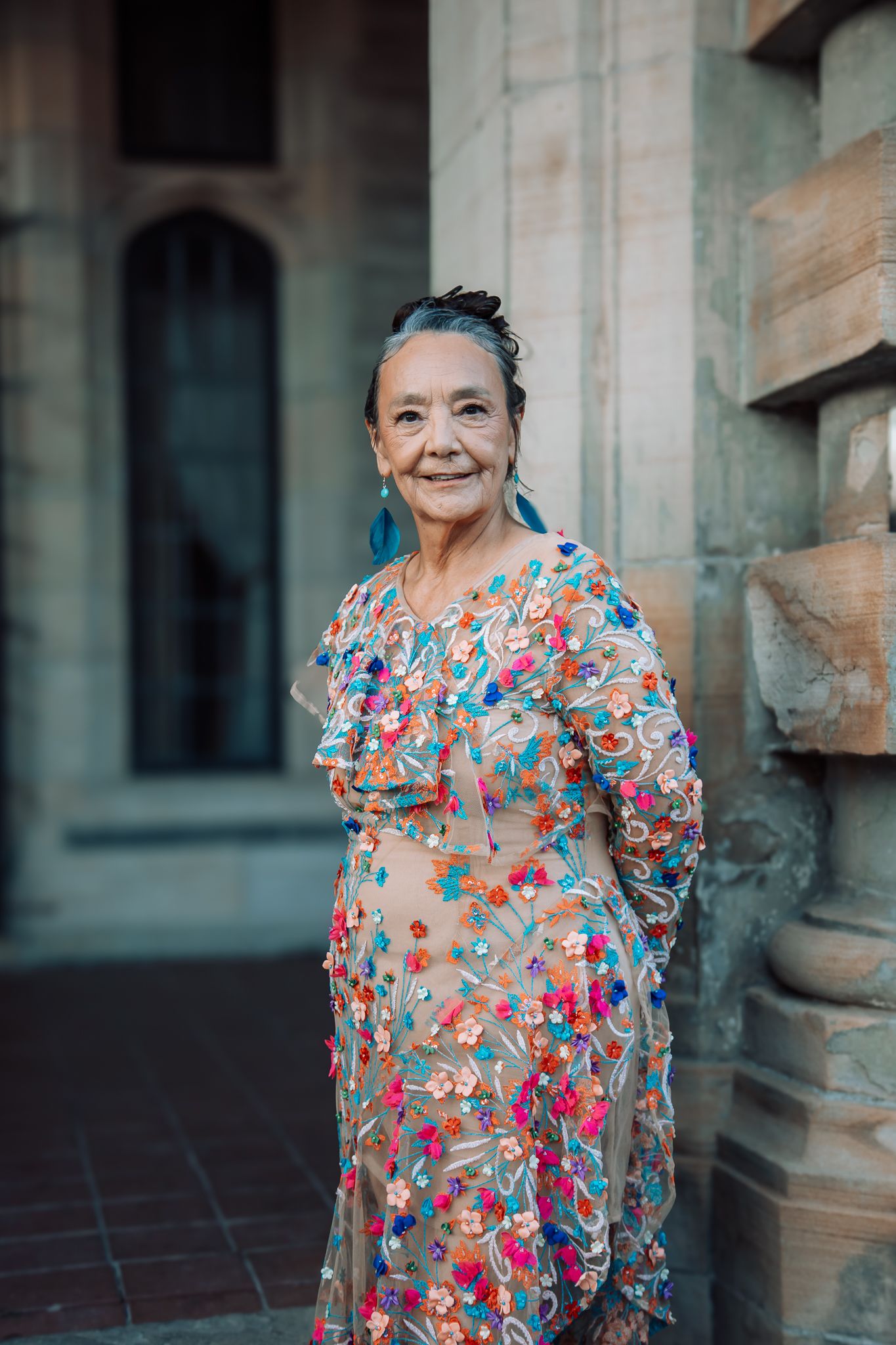 Elderly woman smiling in colorful floral embroidered sheer dress leaning against stone column.