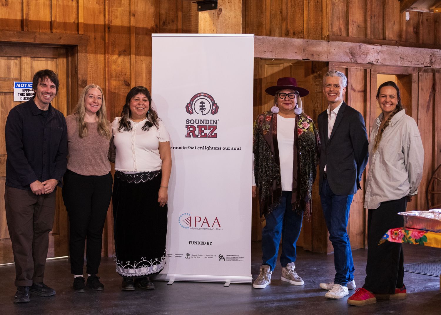 Six people pose together beside a Soundin Rez and IPAA event banner inside a wooden venue.