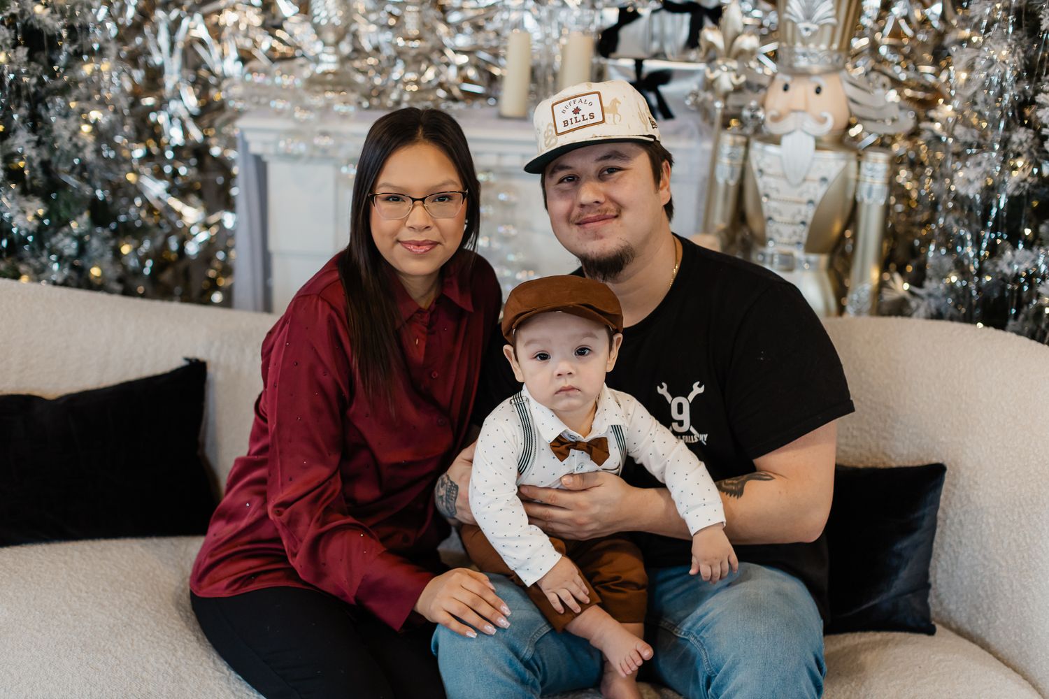 Family of three posing in front of Christmas trees, parents sitting with baby dressed in bow tie and newsboy cap.