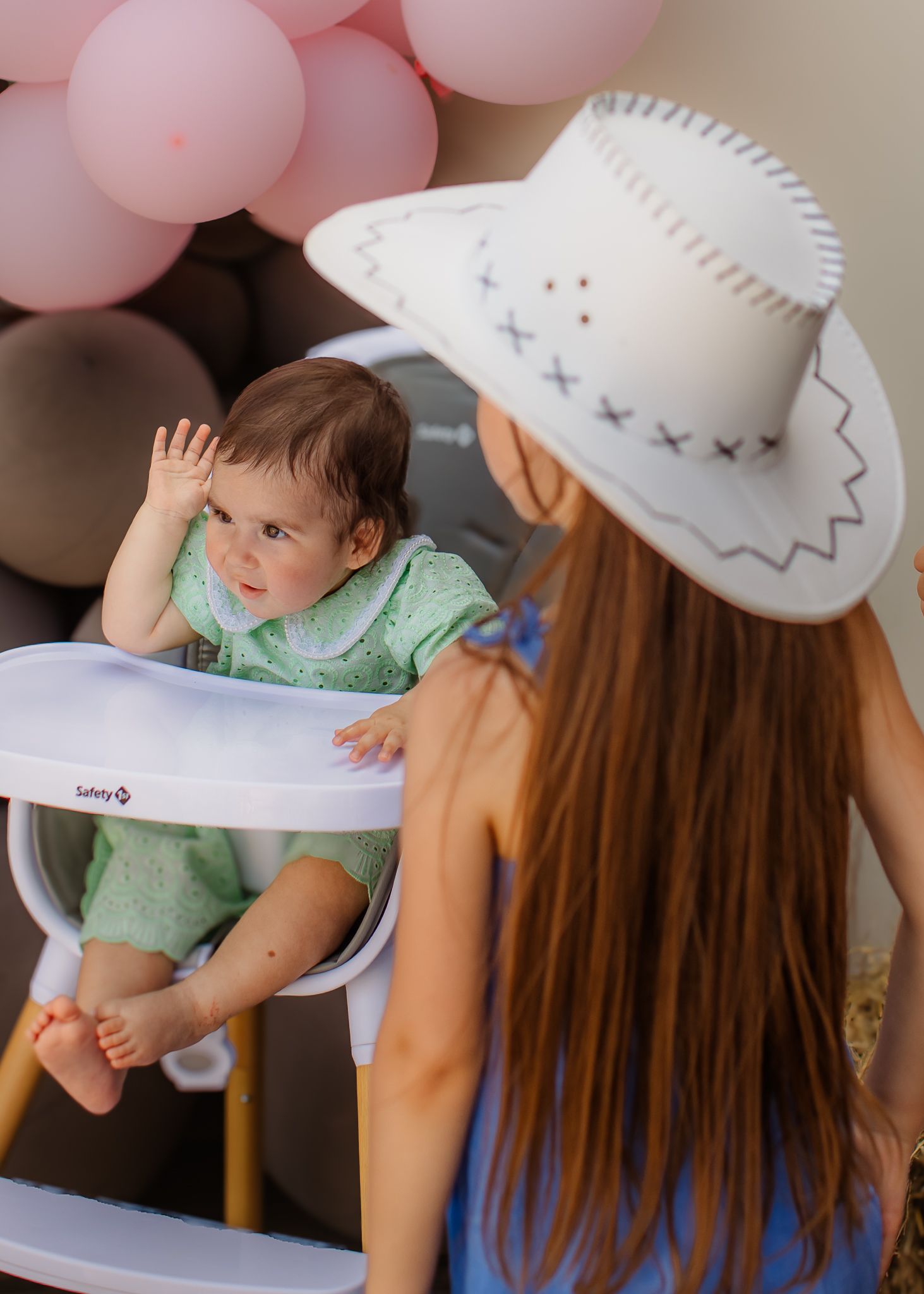 Baby in a Safety 1st high chair at a pink balloon party, with a woman in a white cowboy hat facing away.