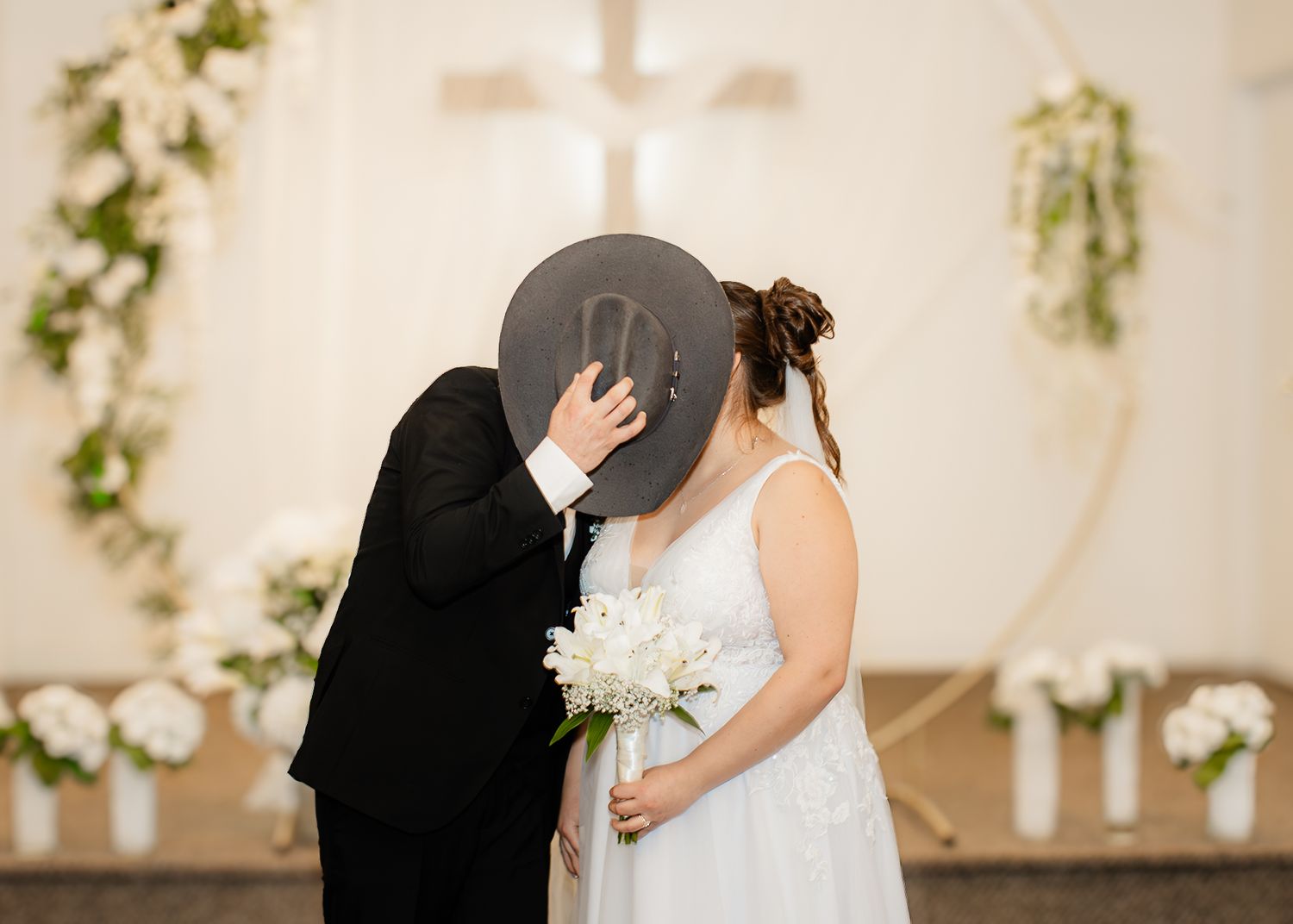 Bride and groom share a kiss hidden behind a cowboy hat at a church wedding ceremony with floral arch.