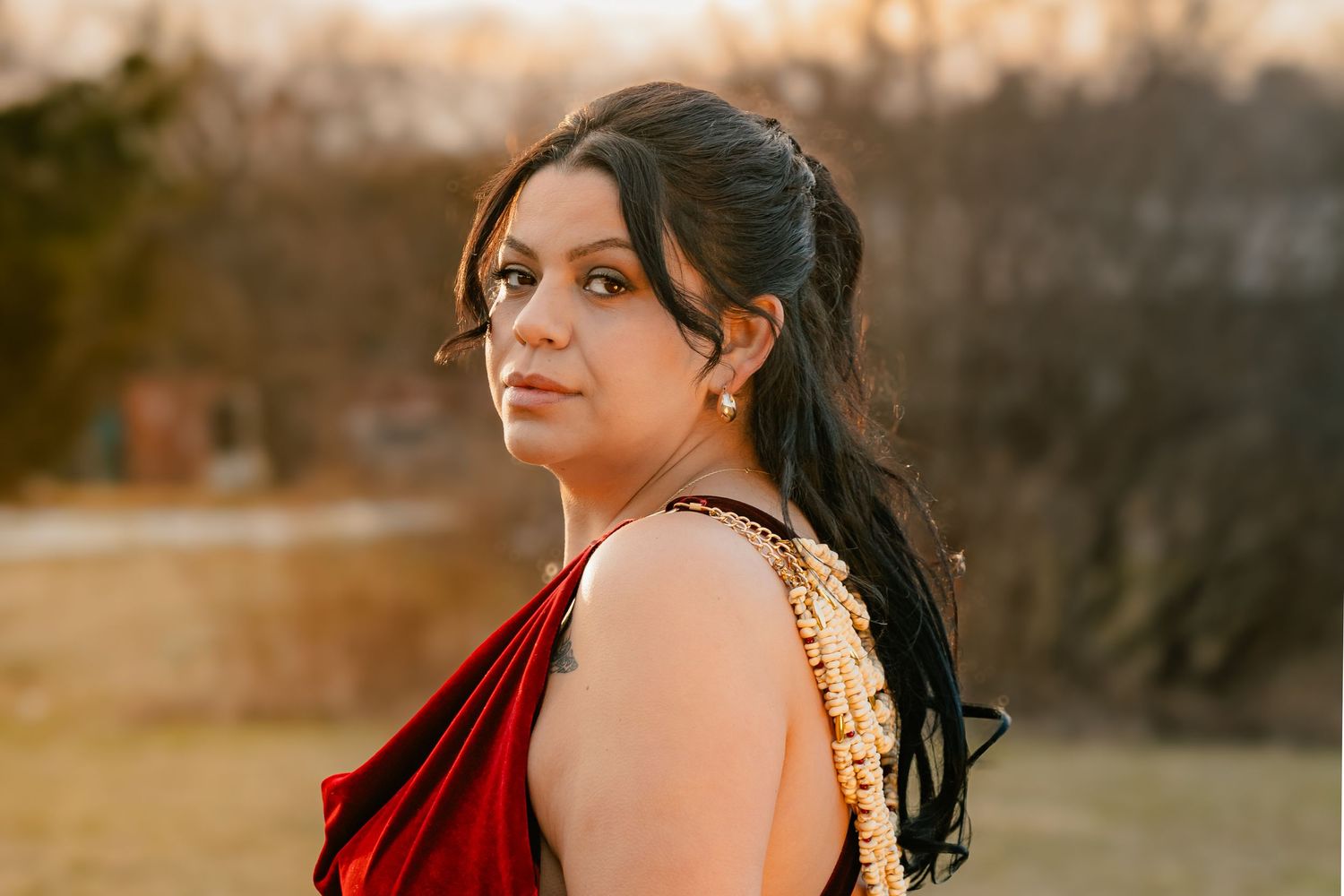 Woman in red velvet dress with gold accents poses outdoors at golden hour with hair styled elegantly.