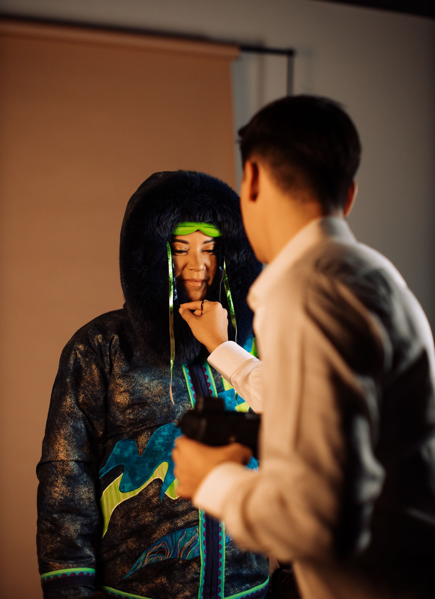 A person in a black hoodie with neon green accents and braids stands while another adjusts their outfit indoors.