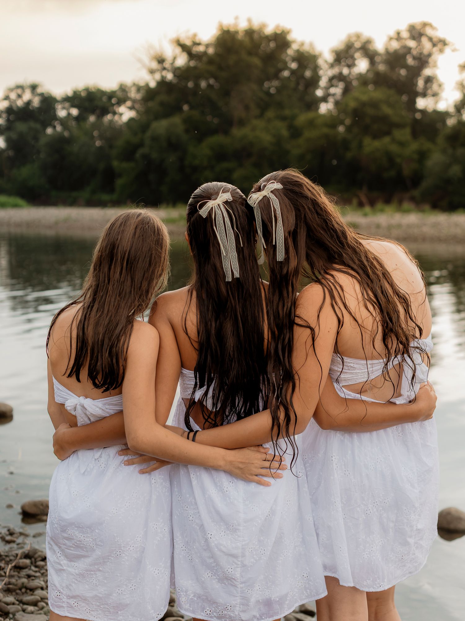 Three girls in white dresses with hair ribbons embrace by a serene lakeside at dusk, viewed from behind.