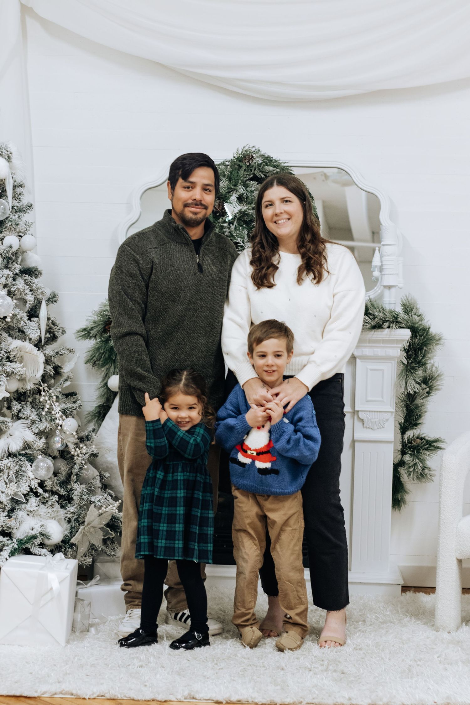 Family holiday portrait in front of white fireplace with snow-covered Christmas trees.