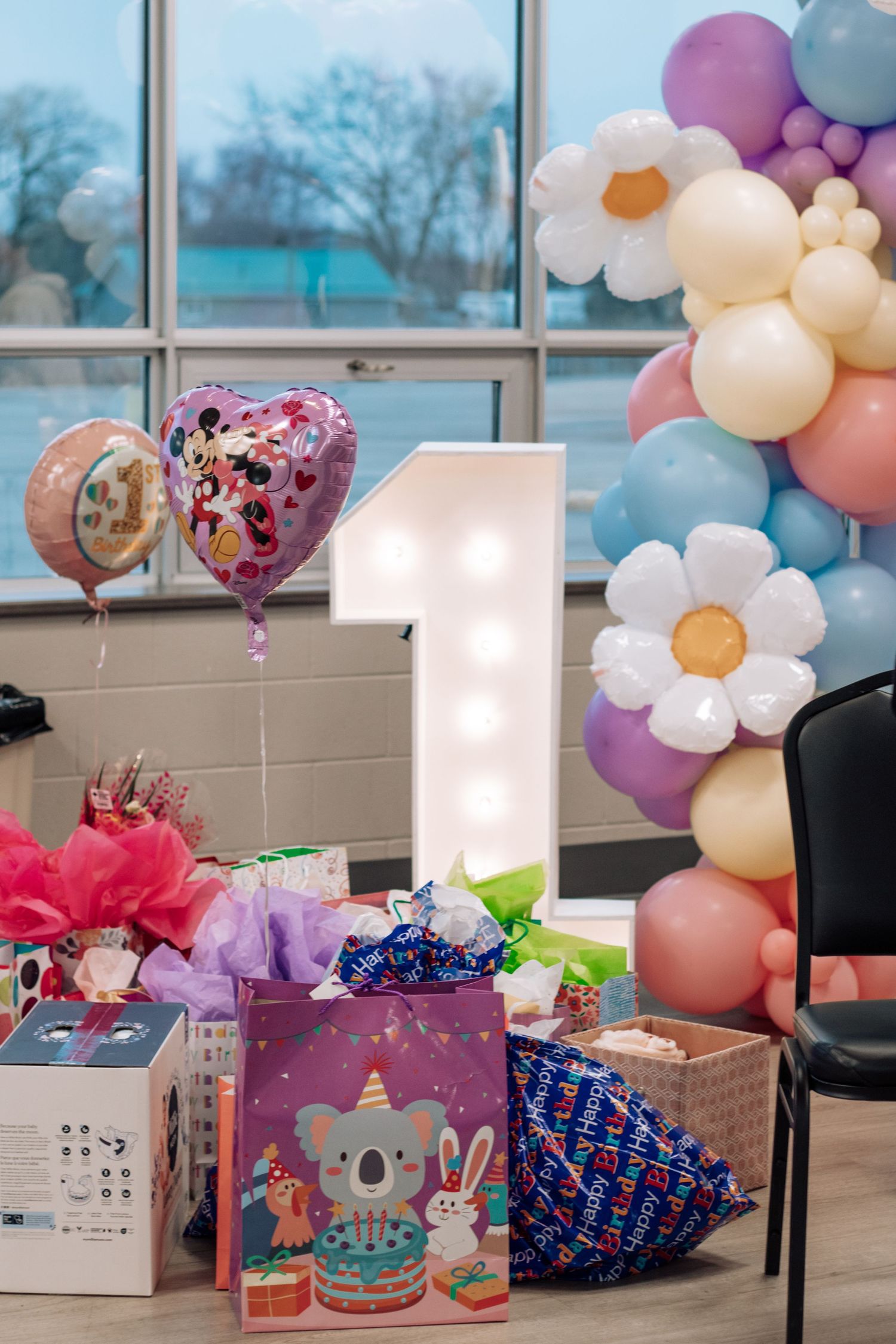 Party setup with pastel balloon garland and gift displays near large windows.